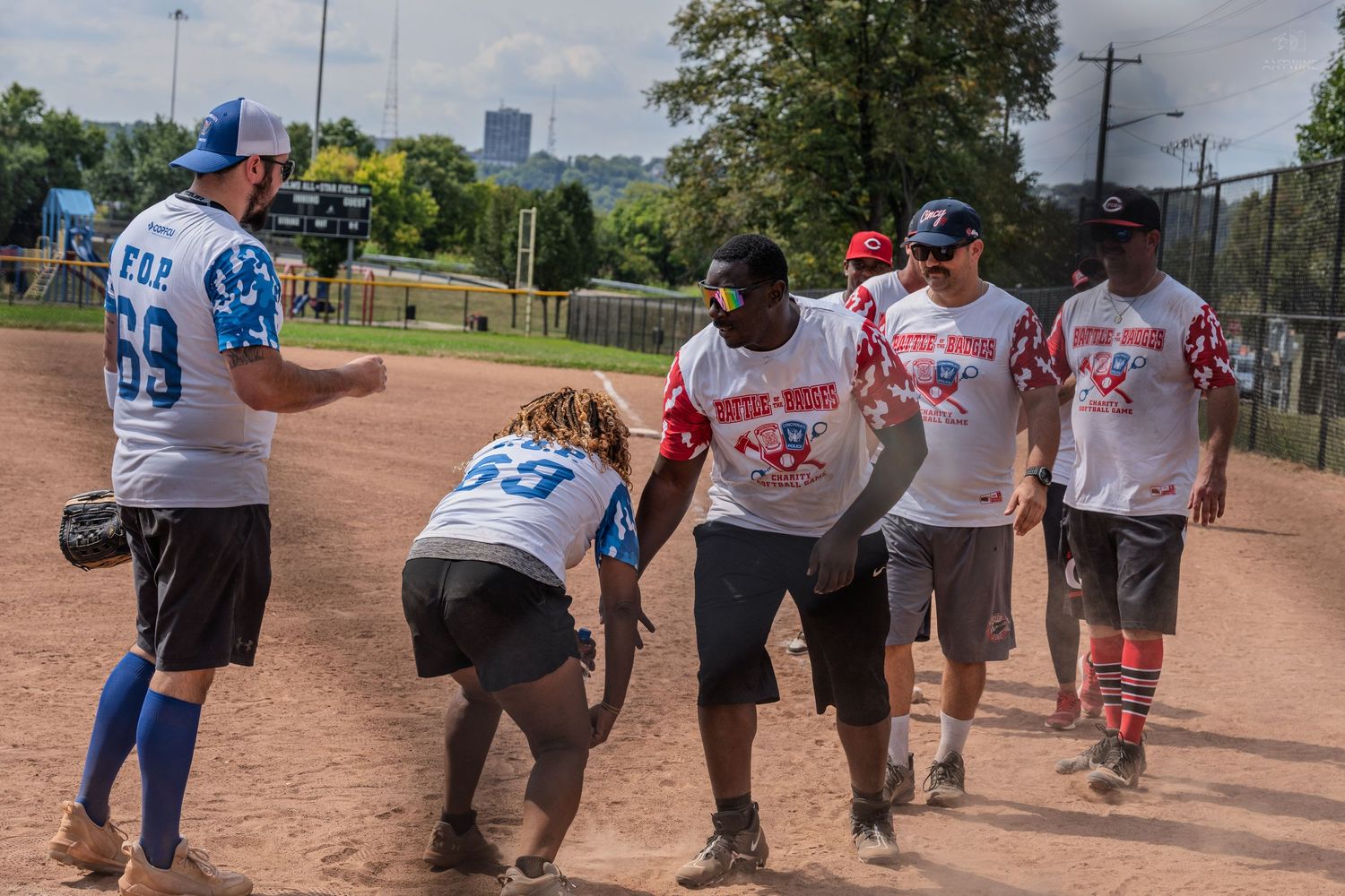 Teams interact on a dirt baseball field during a Battle of the Badges charity softball game on a sunny day.