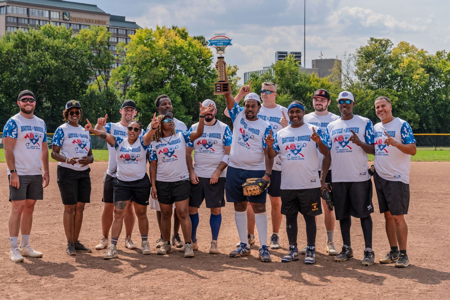 A team in matching Bottle vs Badges jerseys poses together holding a trophy on a softball field.