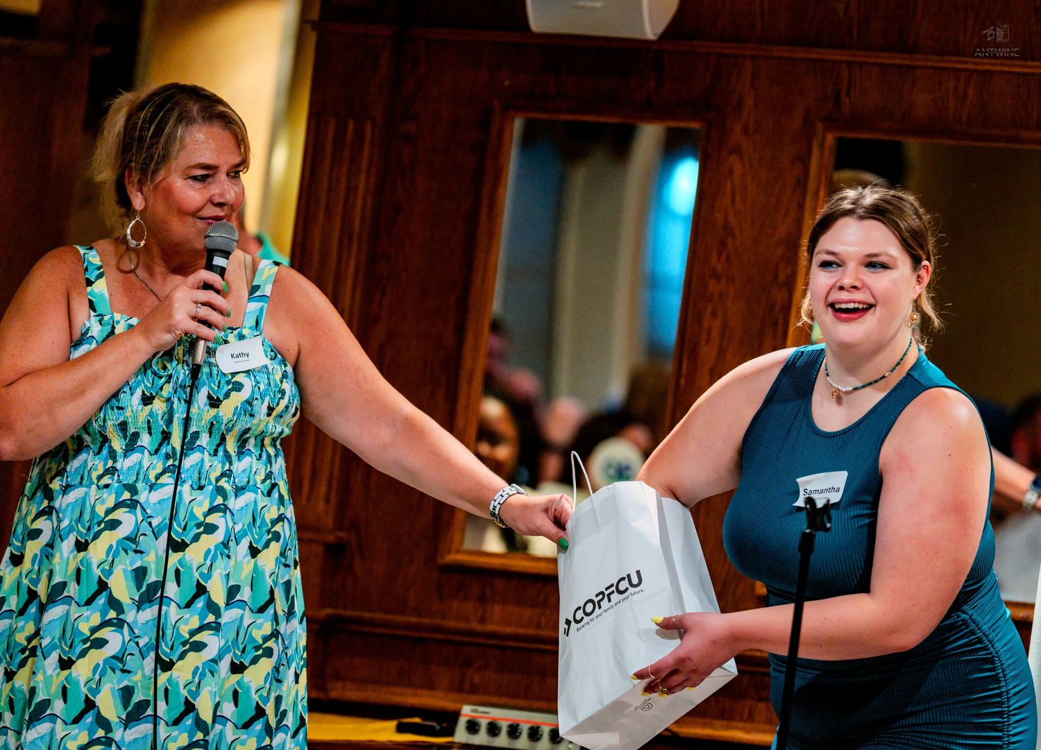 Woman in floral dress presents gift bag to smiling woman in teal at indoor event.