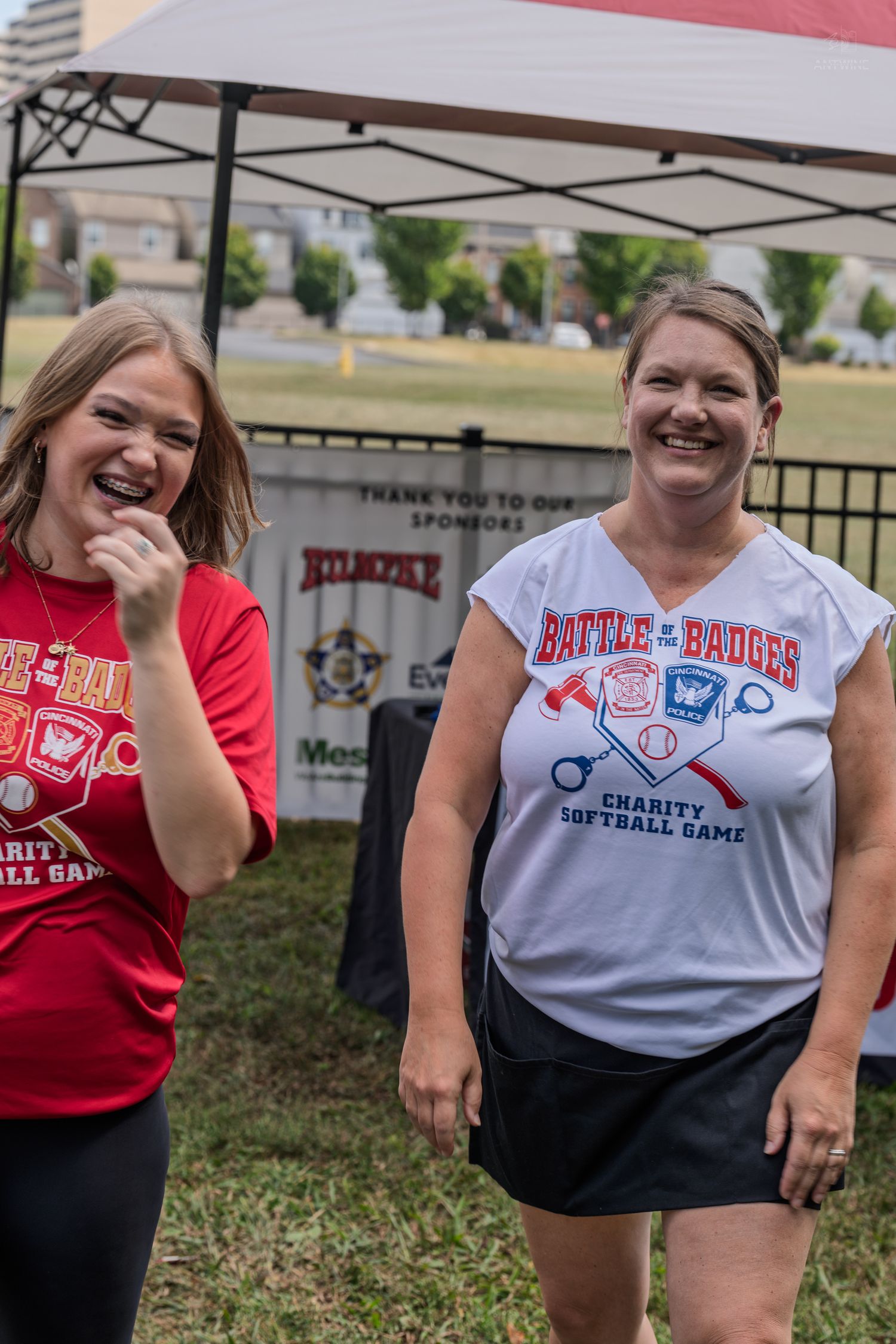 Two women laugh together at a Battle of the Badges charity softball game event outdoors.