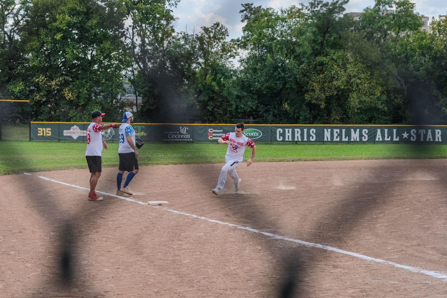 Softball players running bases at Chris Nelms All-Star charity game on a sunny day.