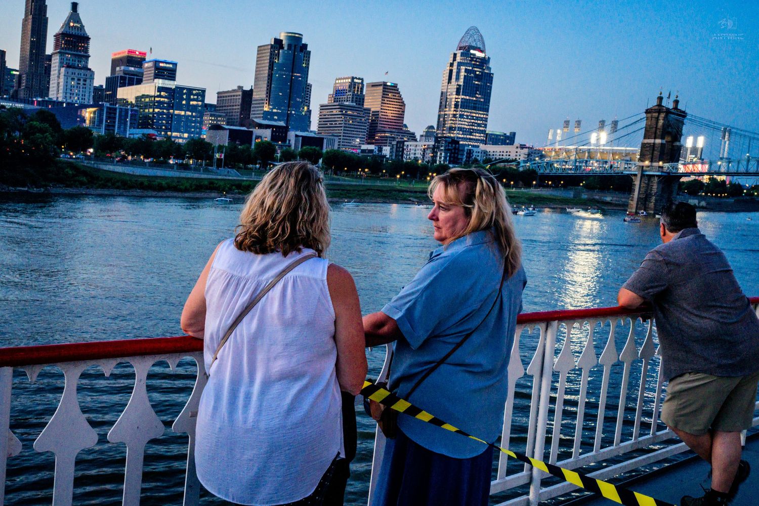 Three people lean on a railing of a riverboat admiring a city skyline at dusk.