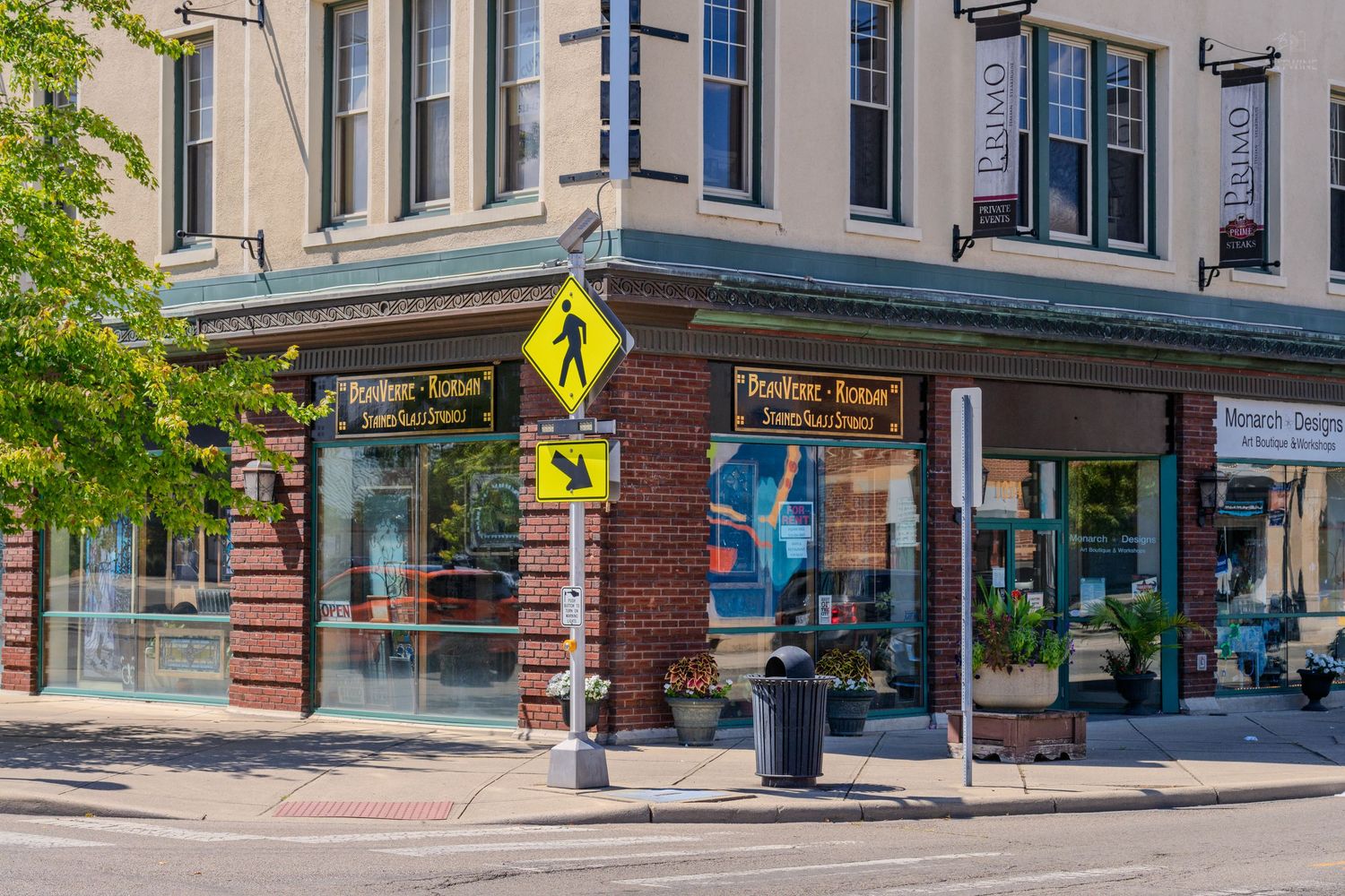 Corner brick building with Bonjour Ketan store, pedestrian crossing sign, and storefronts on a sunny day.