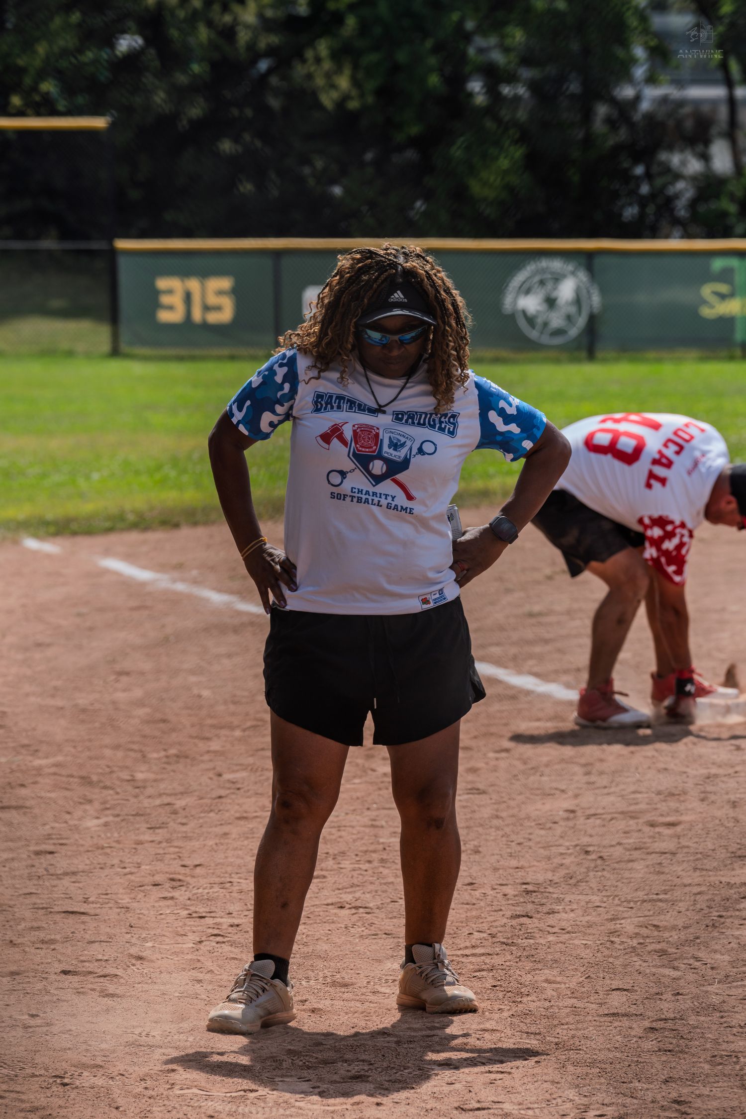 Woman in softball jersey stands on baseball diamond with hands on hips during a sunny outdoor game.