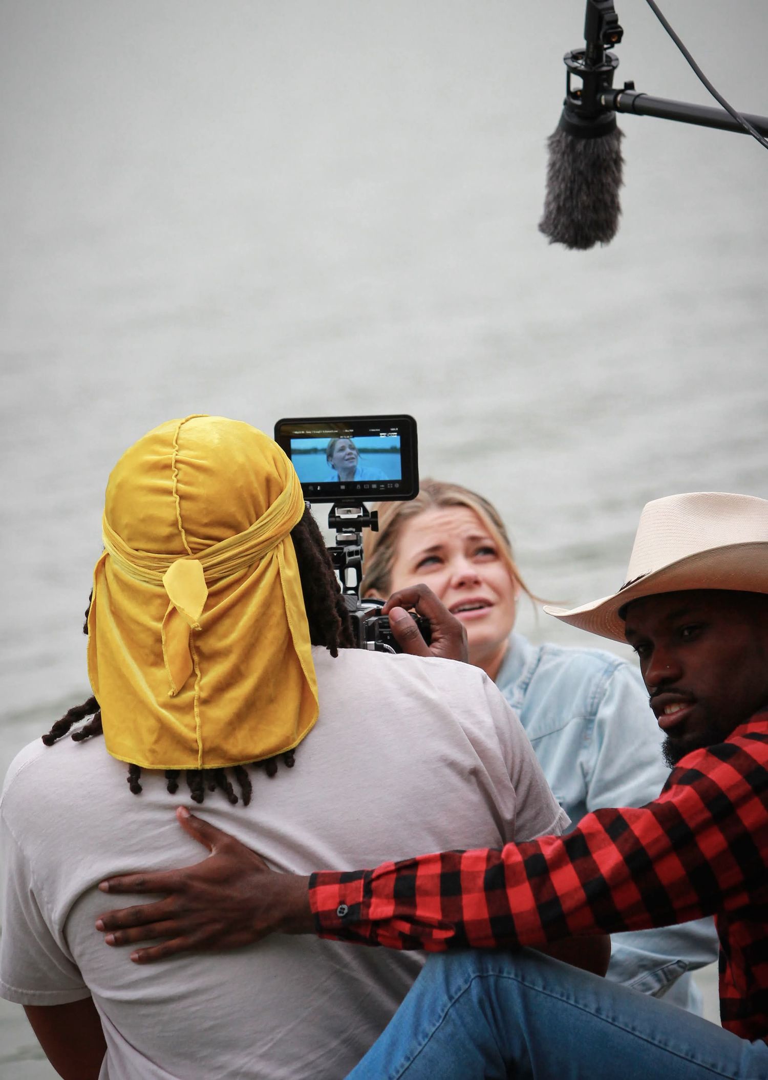 Film crew capturing emotional scene outdoors near water, with boom mic, camera monitor, and actors embracing.