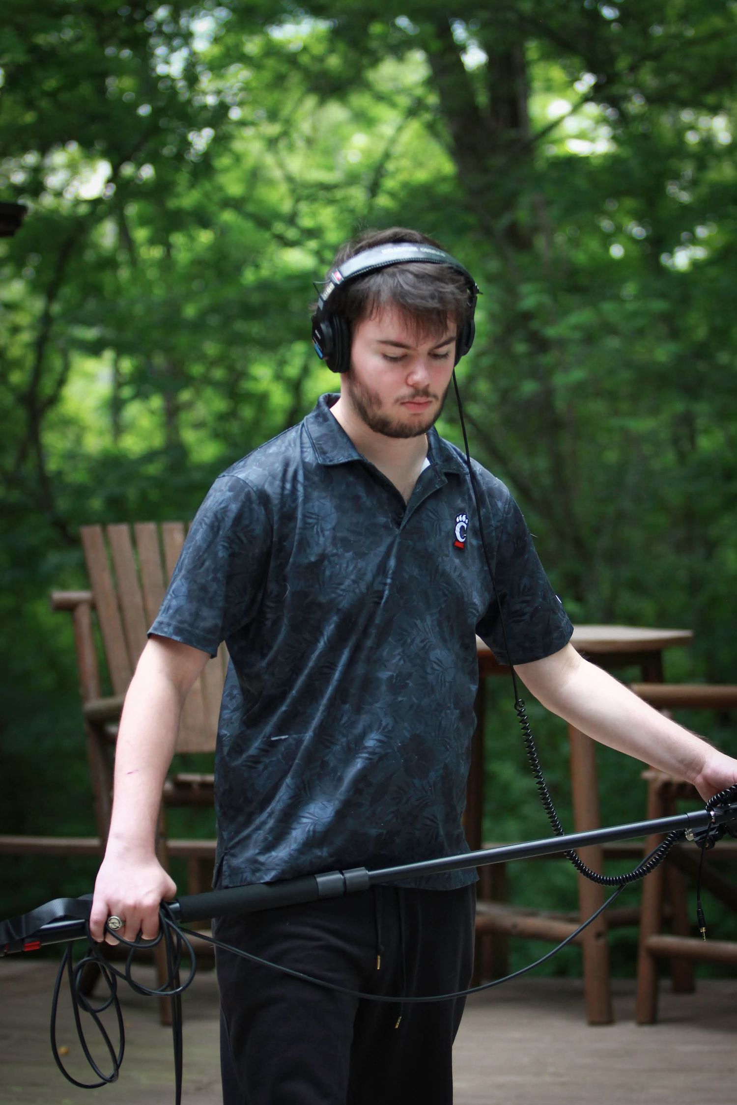 Young man with headphones holds a boom microphone on a wooden deck surrounded by lush green trees.
