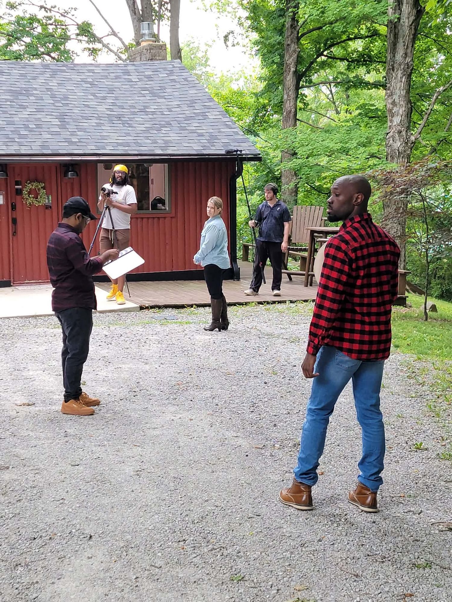 Film crew rehearsing outside red cabin in wooded area, actors reviewing scripts on gravel path.