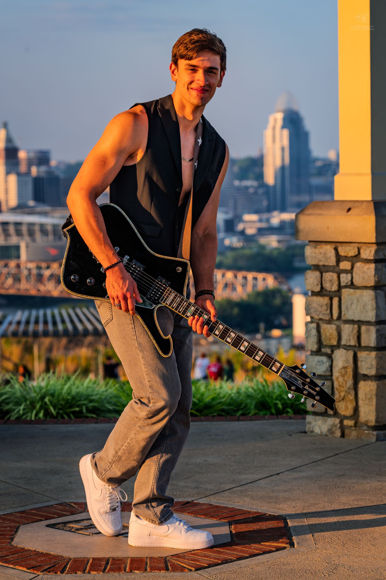 Young man playing black electric guitar outdoors at golden hour with city skyline in background.