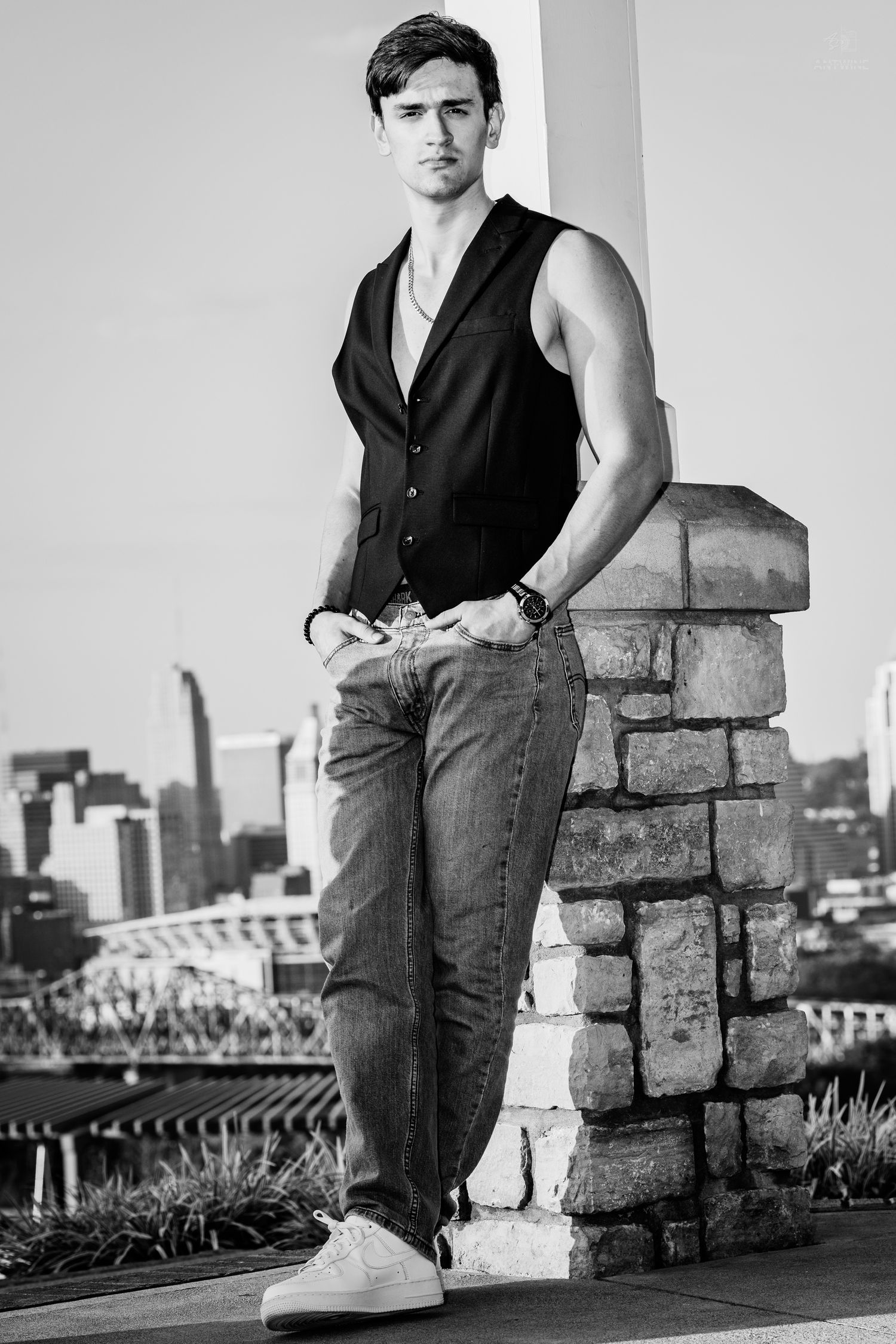 Young man in sleeveless vest and jeans leaning against stone pillar with city skyline in black and white.