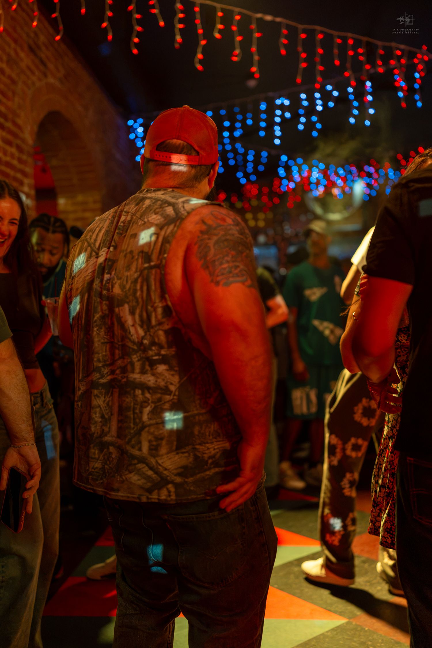Man with tattoos wearing a red cap and camo vest at a vibrant nightclub with colorful string lights.