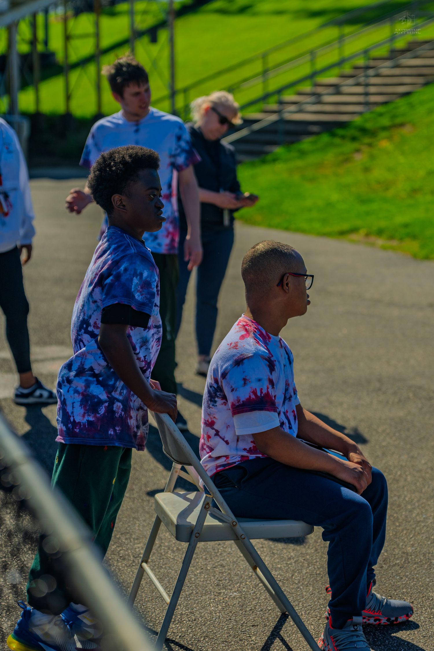 Youth athletes in tie-dye shirts gather near bleachers on a sunny day, one seated in a folding chair.