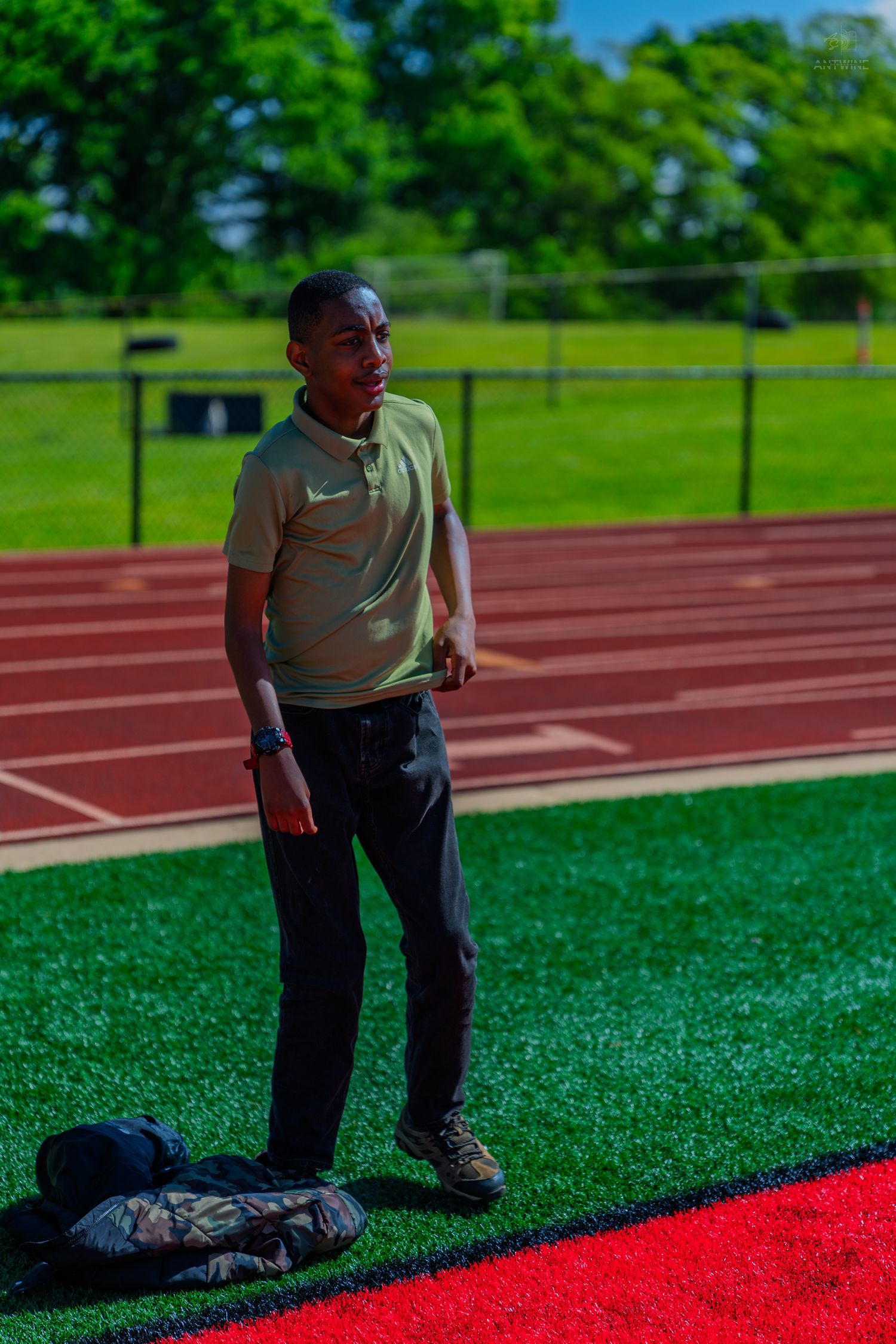 Young athlete standing on green turf beside a red running track on a sunny day.