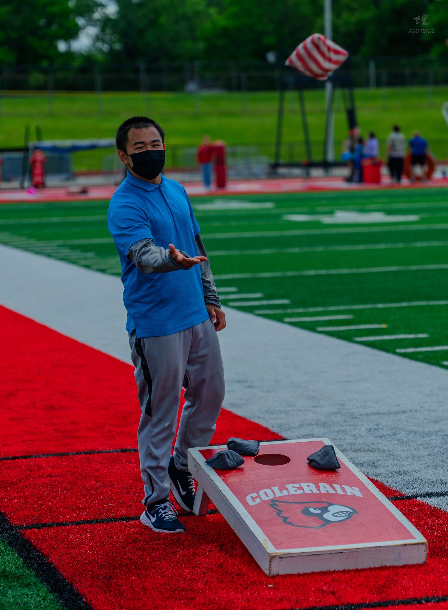 A masked boy plays cornhole on a red turf field next to a Colerain branded board outdoors.