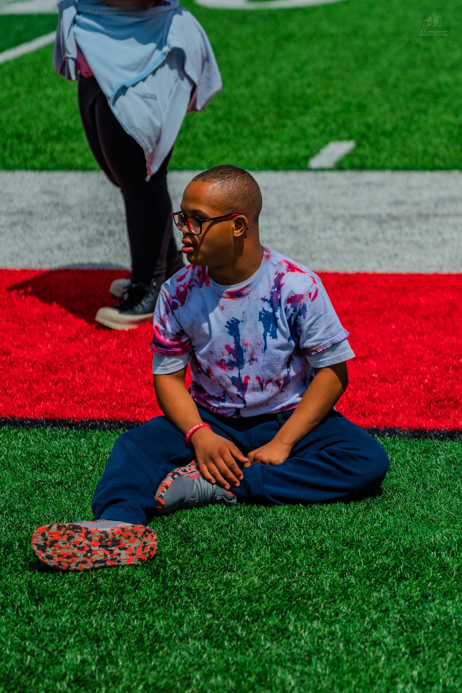 Young boy with glasses sits cross-legged on green turf field wearing tie-dye shirt and navy sweatpants.