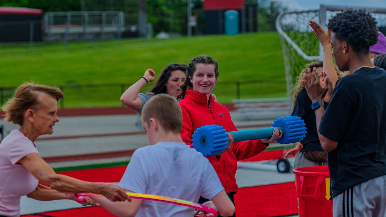 Group of people enjoying an outdoor track event, engaging in fun activities with foam noodles on a red track.