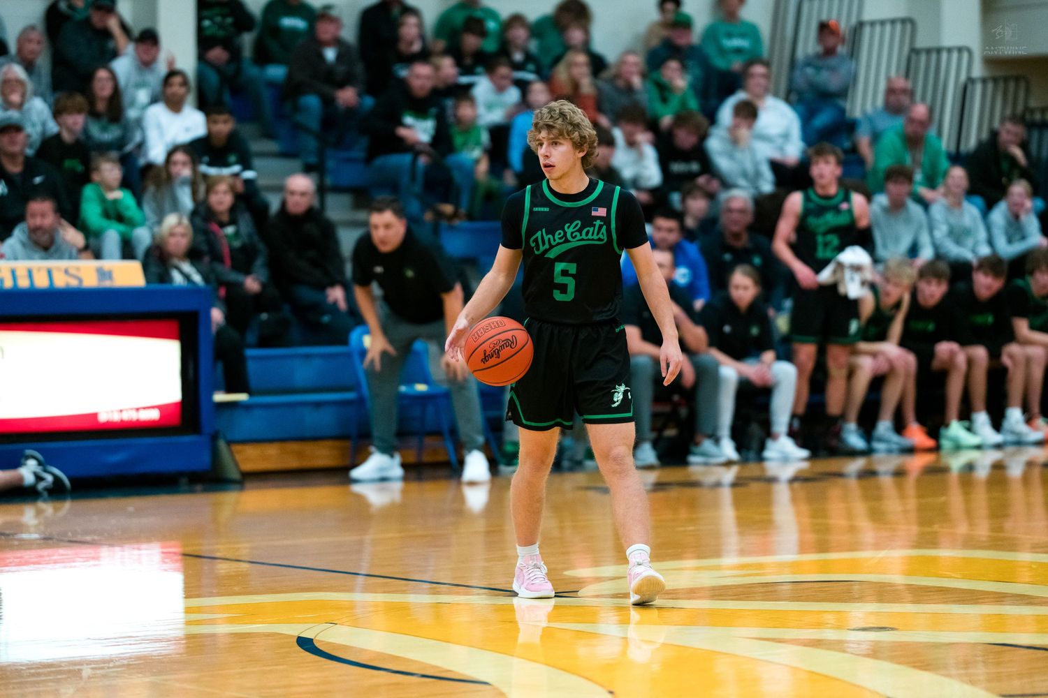 Basketball player wearing green number 5 jersey dribbles ball on hardwood court during game.