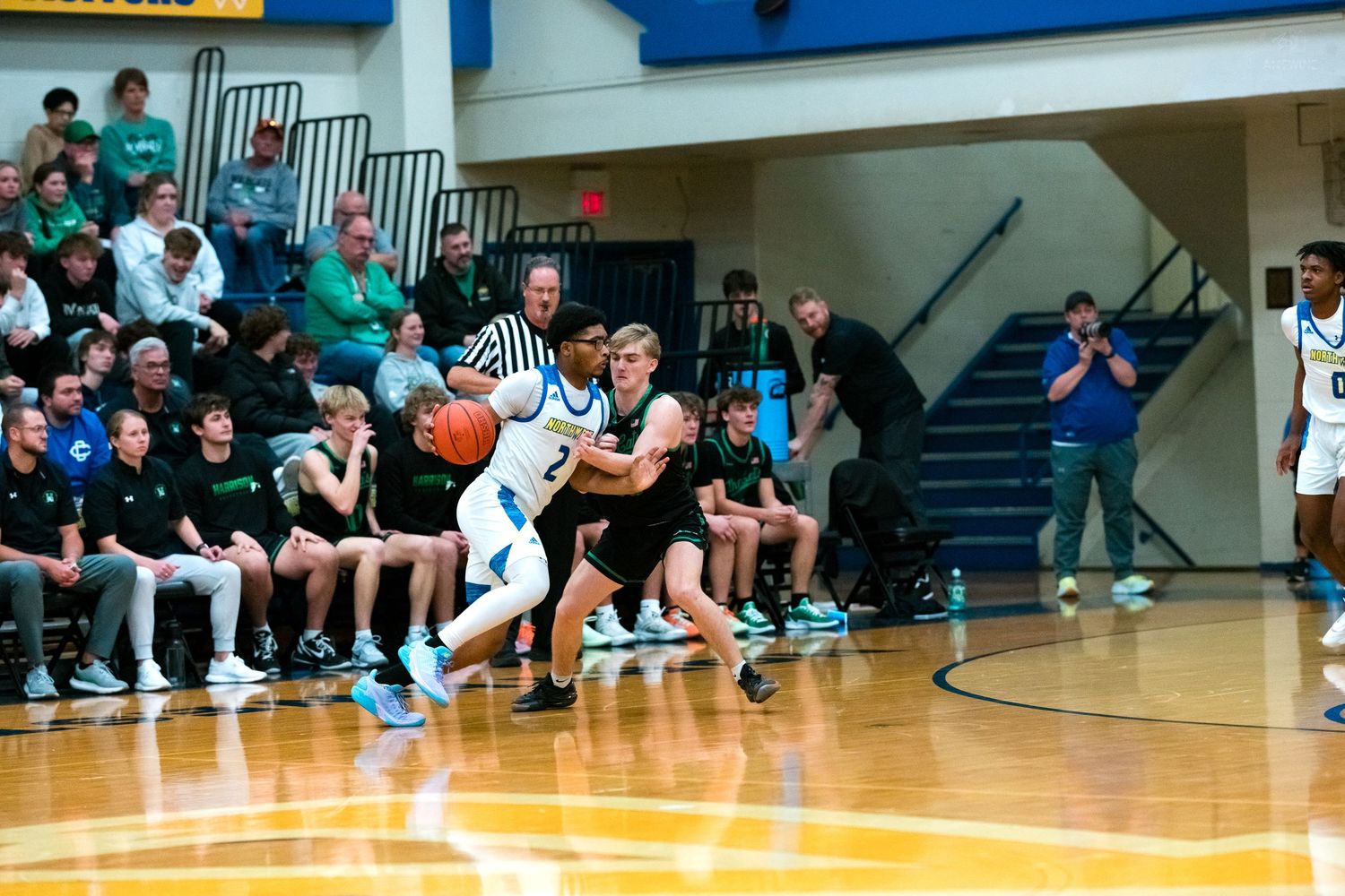 High school basketball player in blue dribbles past green-uniformed defender during fast-paced game with packed crowd.