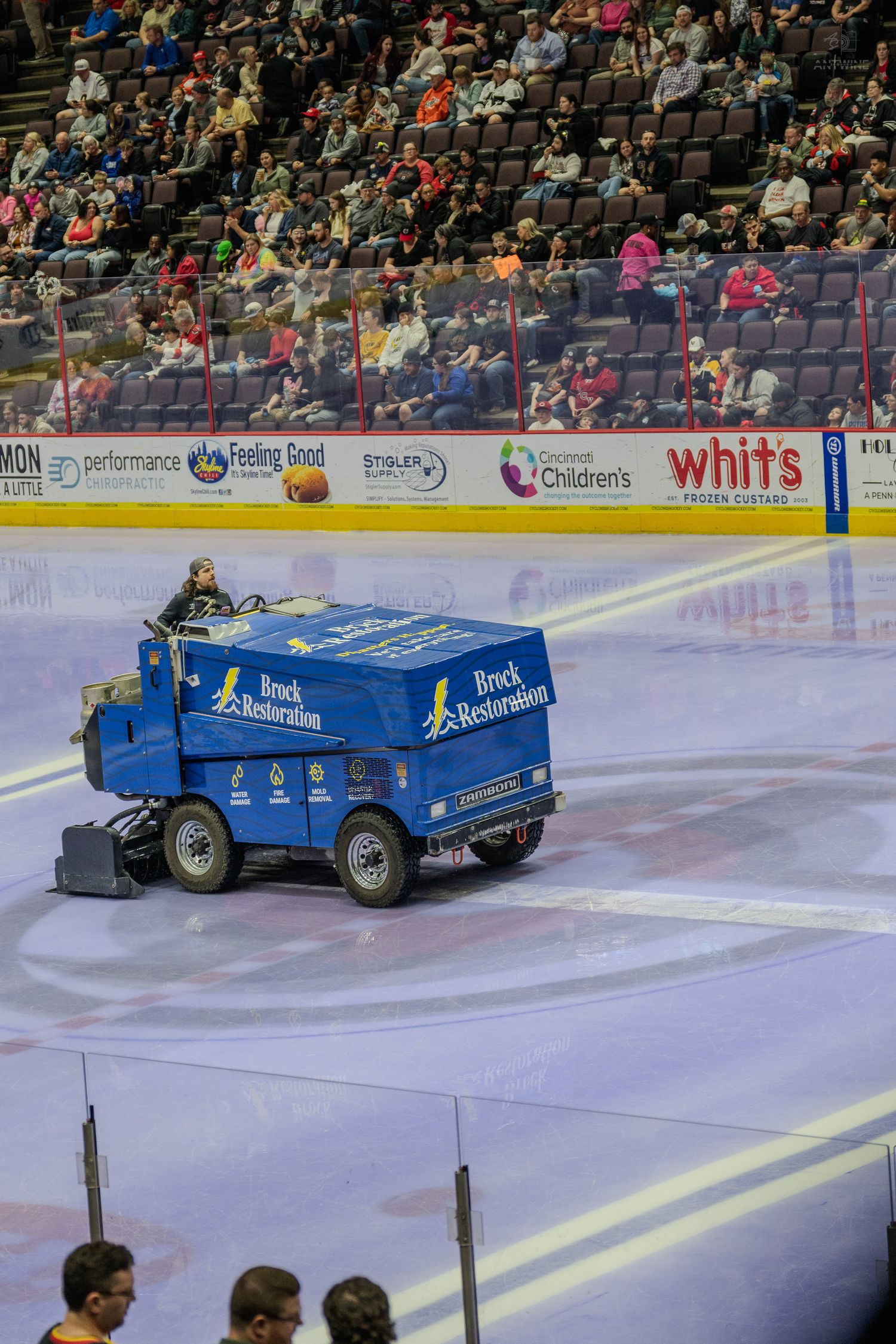 A blue Brock Restoration Zamboni resurfacing the ice at a packed hockey arena during an intermission break.