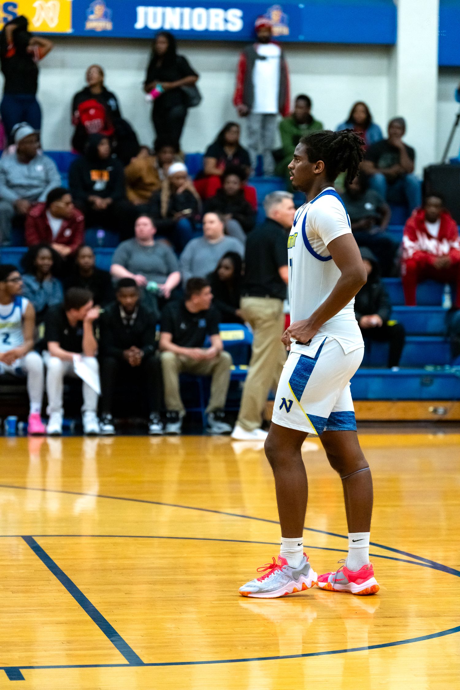 A high school basketball player in a white and blue uniform stands on the court during a game.