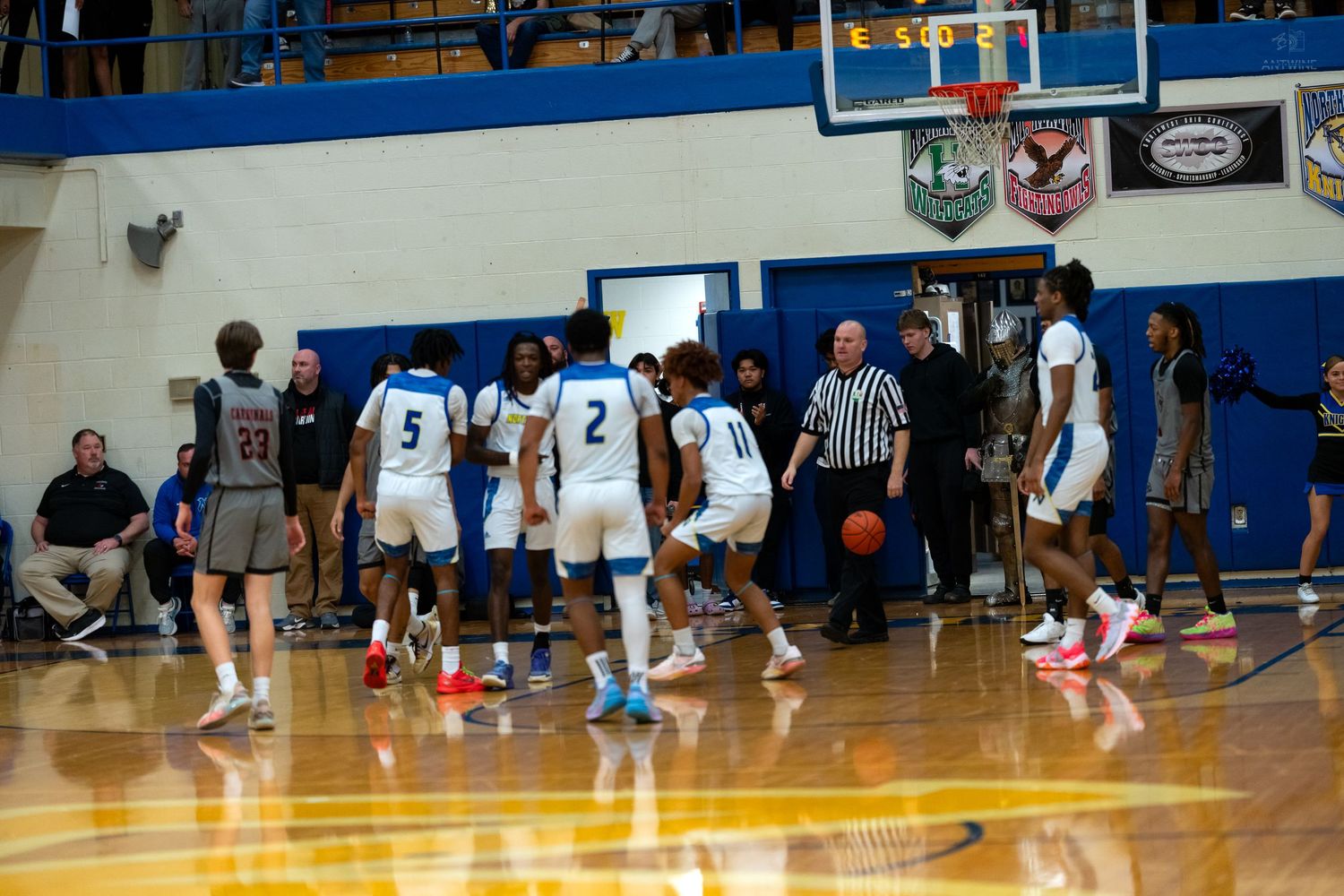 High school basketball players in blue and white uniforms dribble during a game in a decorated school gymnasium.