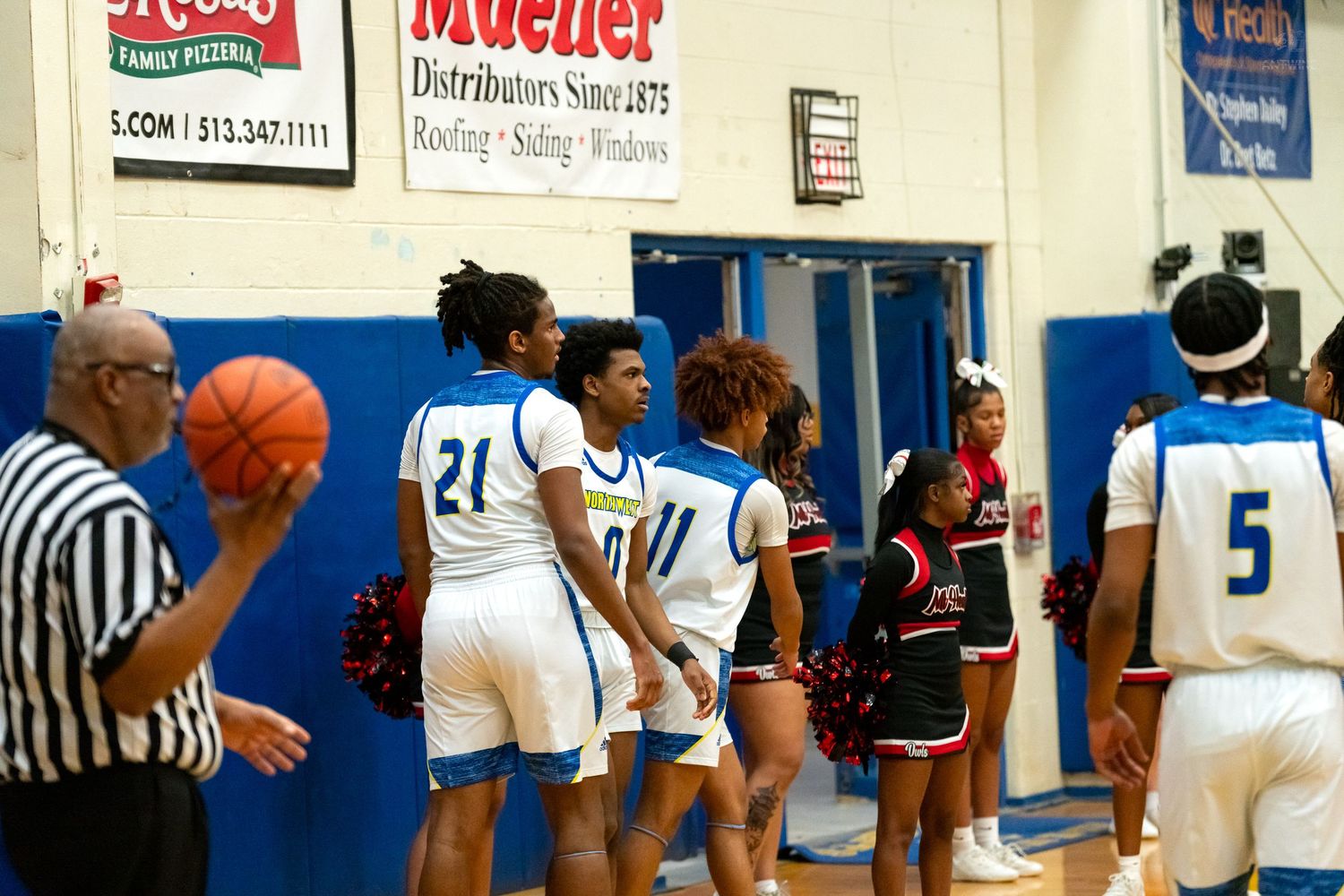 High school basketball players in blue and white uniforms line up during game with cheerleaders nearby.
