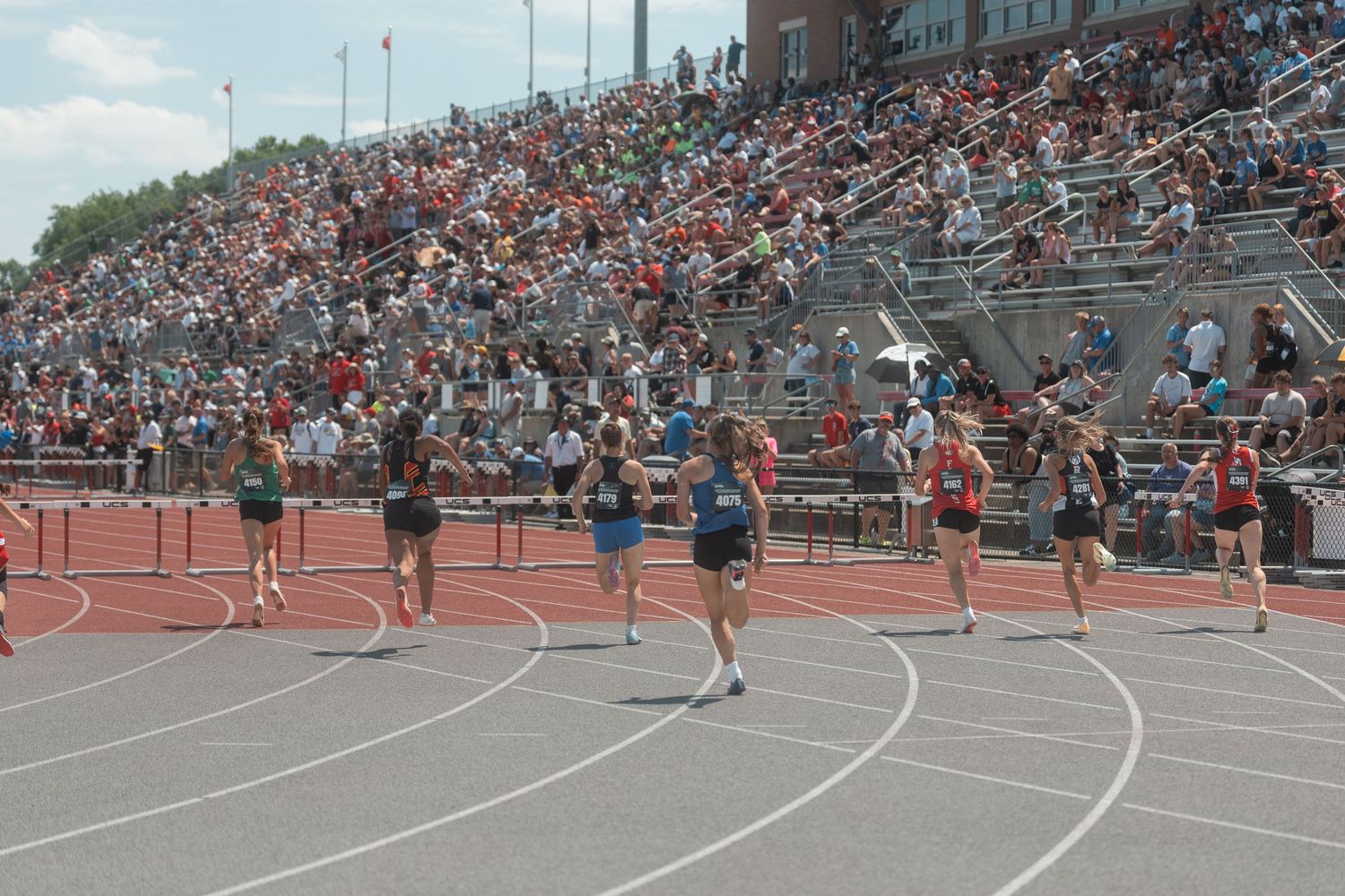 Athletes compete in a hurdles race at a packed track and field stadium during a high school championship meet.