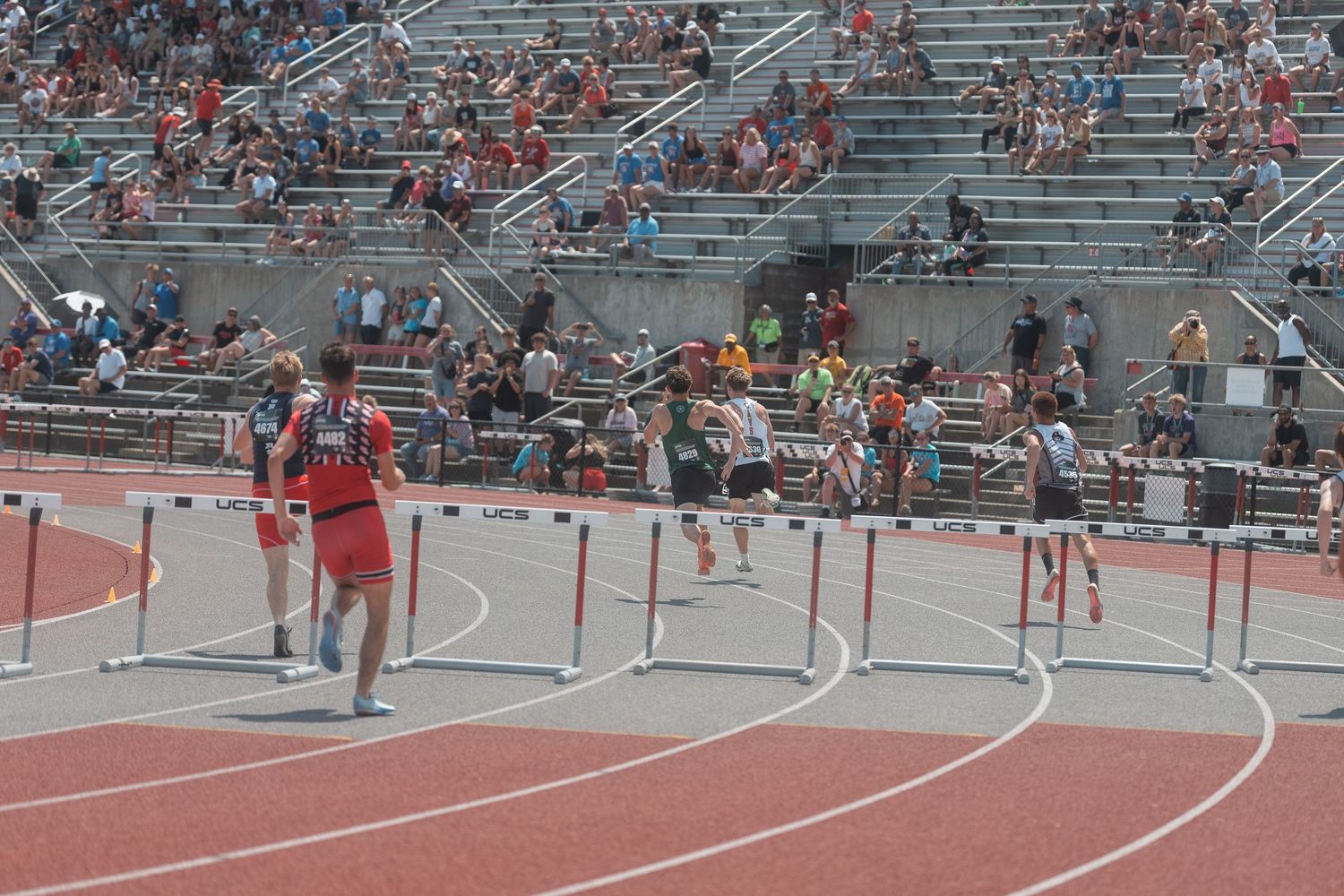 Athletes navigate hurdles on a red track during an outdoor high school track and field competition.