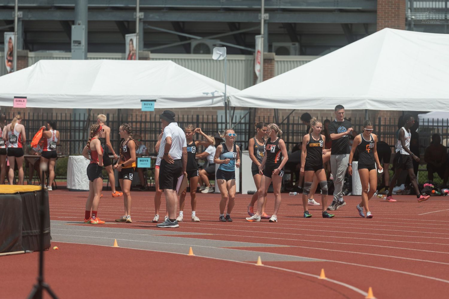 Group of female track and field athletes gather near orange cones on a red track at an outdoor state championship meet.