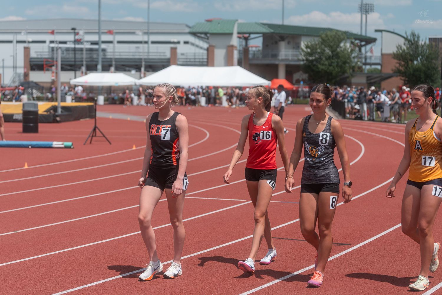 Four female track and field athletes in numbered uniforms walking together on red outdoor competition track.