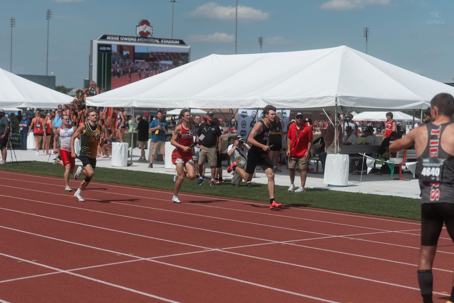 Runners sprint past white tents during relay race at outdoor high school track and field championship event.
