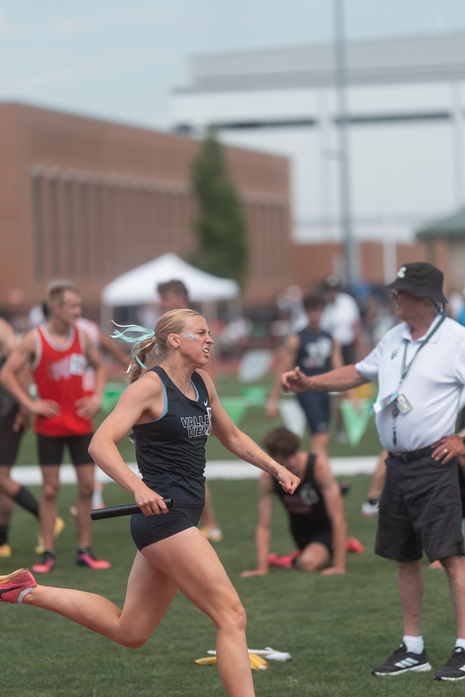 Blonde female athlete runs relay race holding baton at competitive high school track and field event.