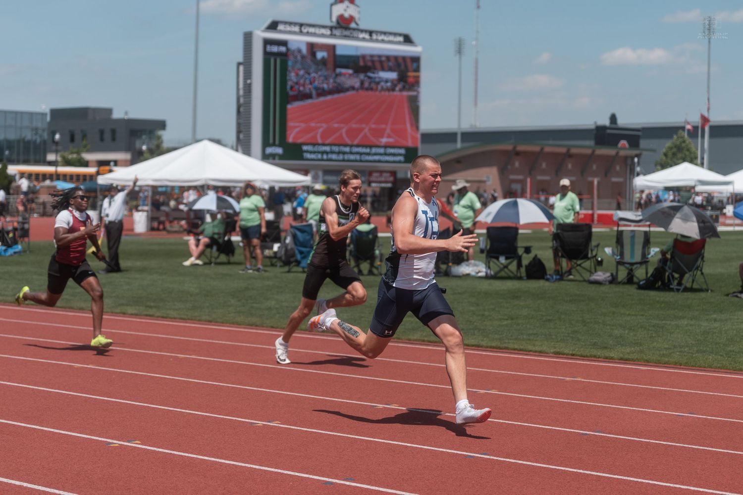 Male athletes sprint on a red track during a competitive meet with a large video scoreboard visible in the background.
