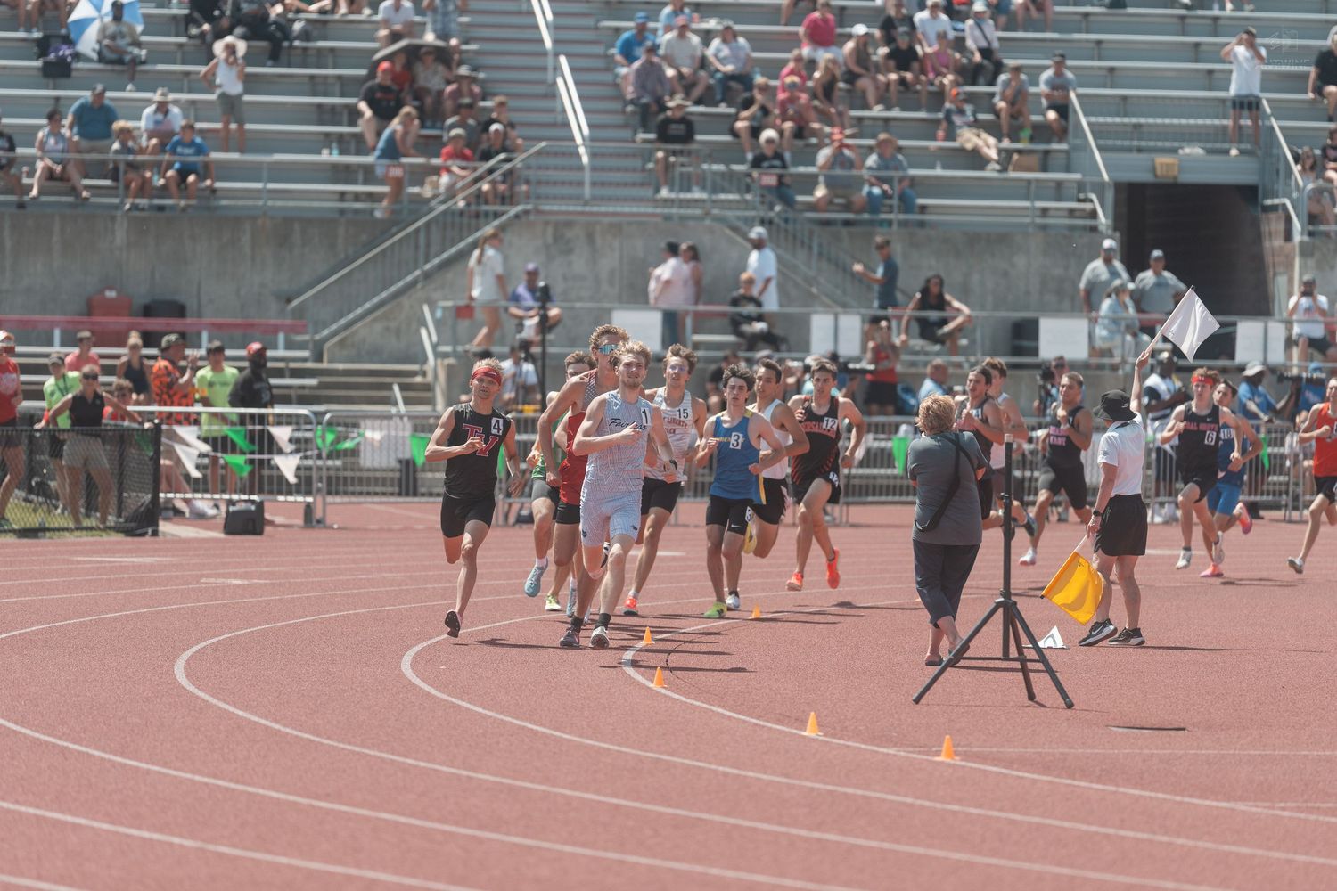 Track and field athletes run on a red track while a cameraman films the race at a sunny meet.