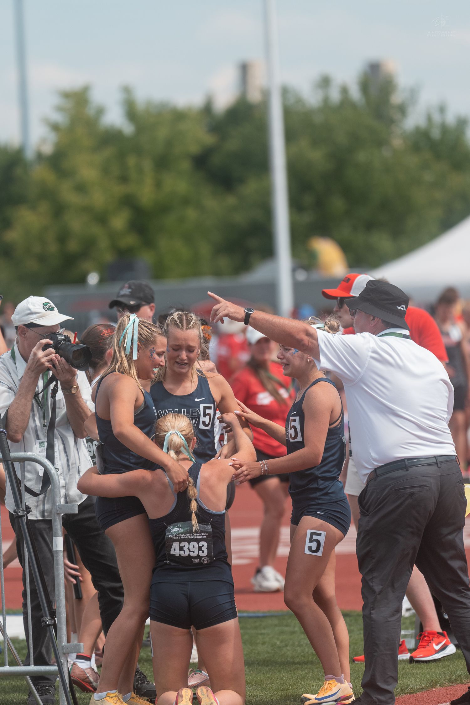 Track and field girls team celebrating with coach pointing directions at a sunny outdoor meet.