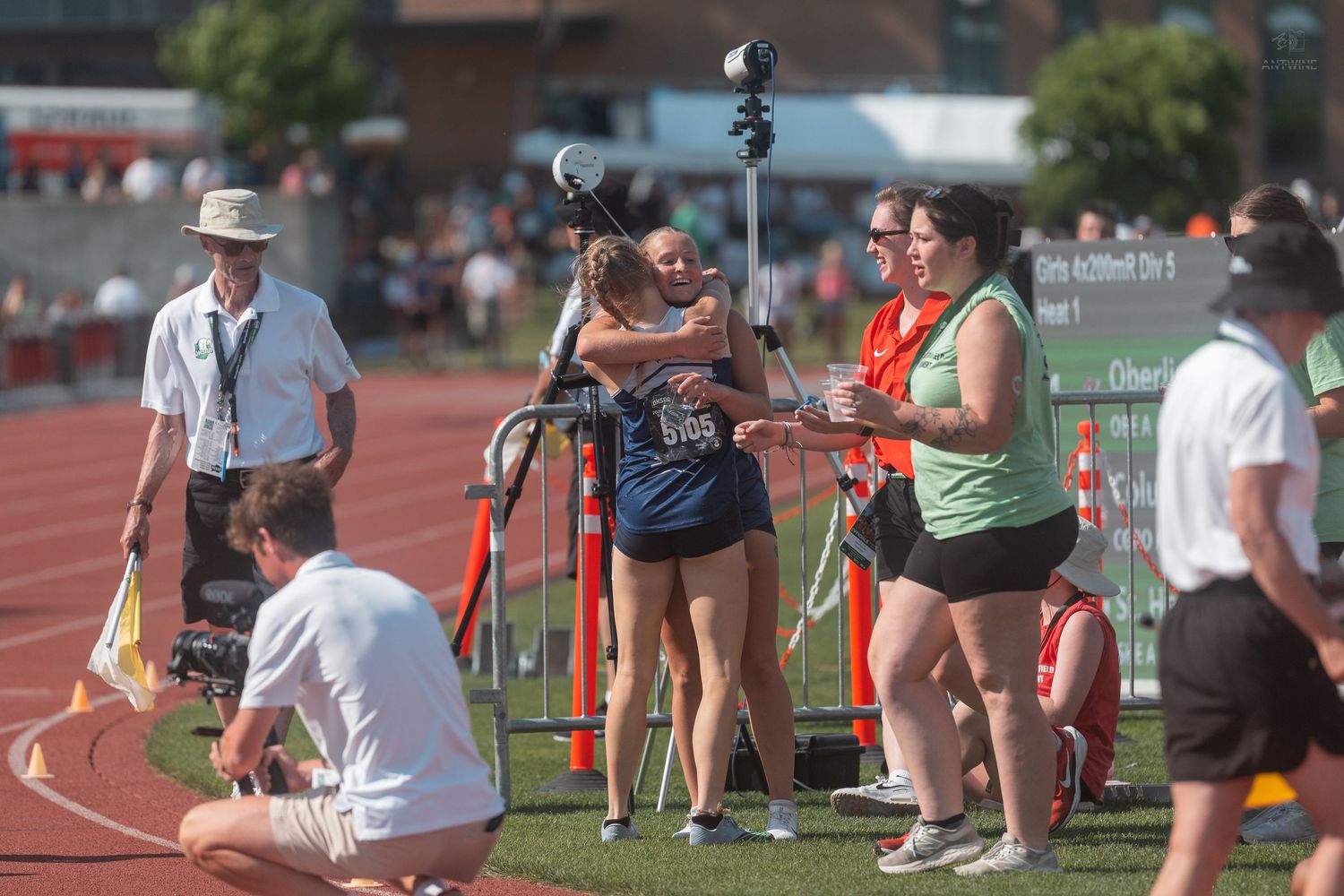 Athletes and coaches celebrate with hugs near the finish line at an outdoor track competition.