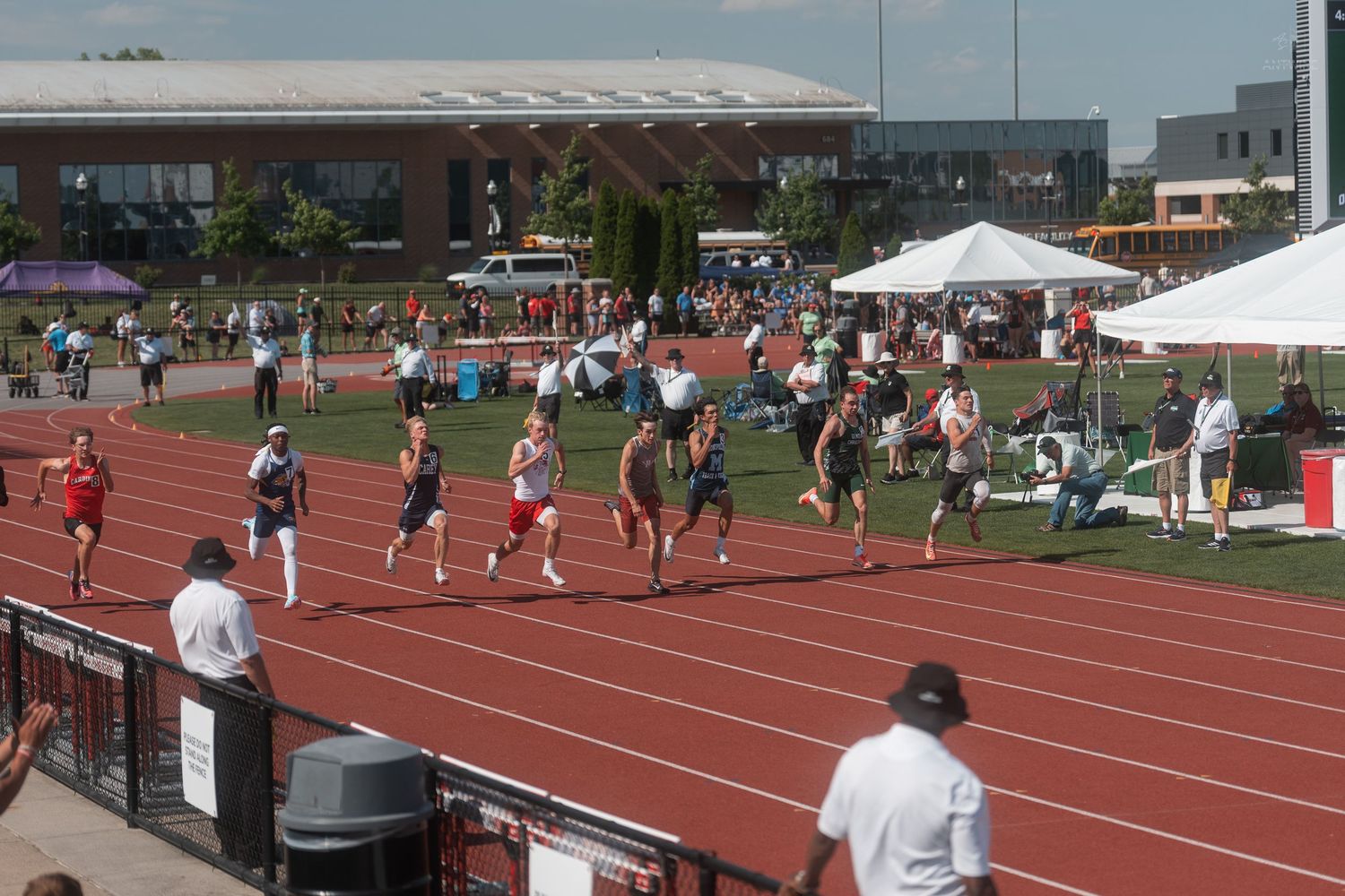 Athletes compete in a track and field race on a red running track during an outdoor meet on a sunny day.