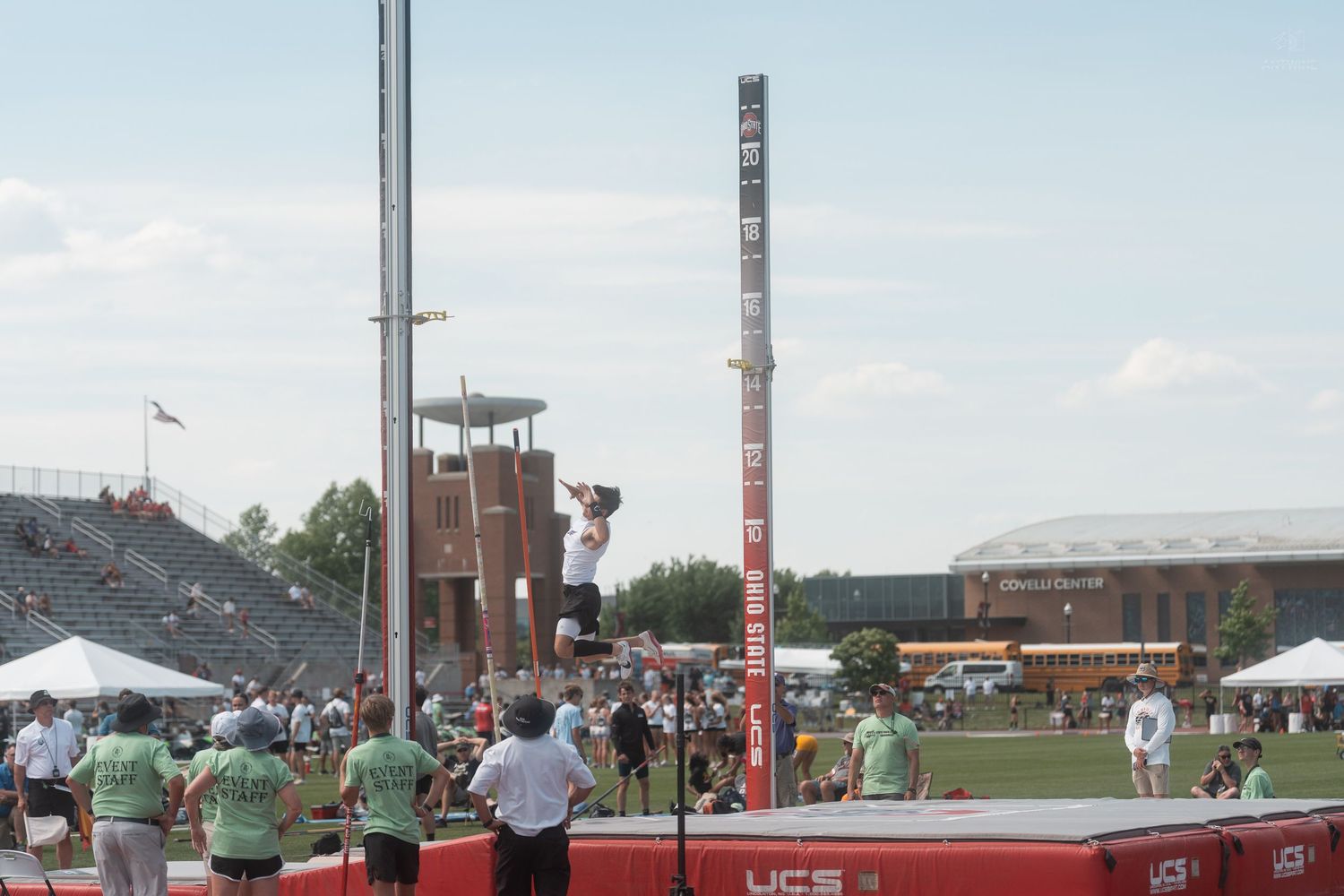 Pole vaulter ascends the pole during competition at an outdoor track and field meet with large stadium crowd.