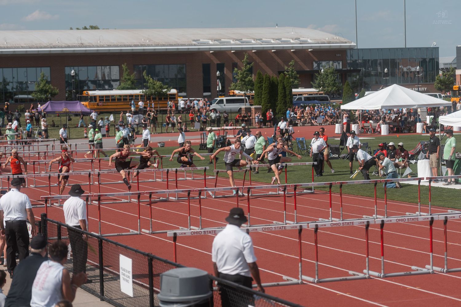 Multiple high school athletes running hurdles race on red track at outdoor championship meet with spectators watching.