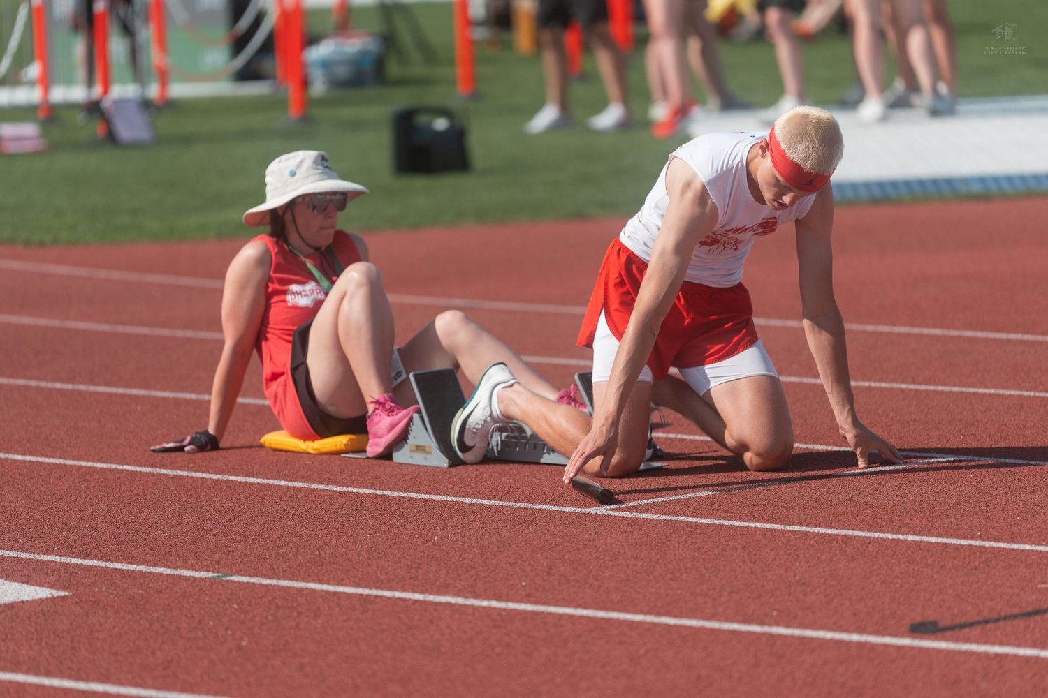 Track athlete in red uniform crouches in starting position on blocks with seated official on sunny day.