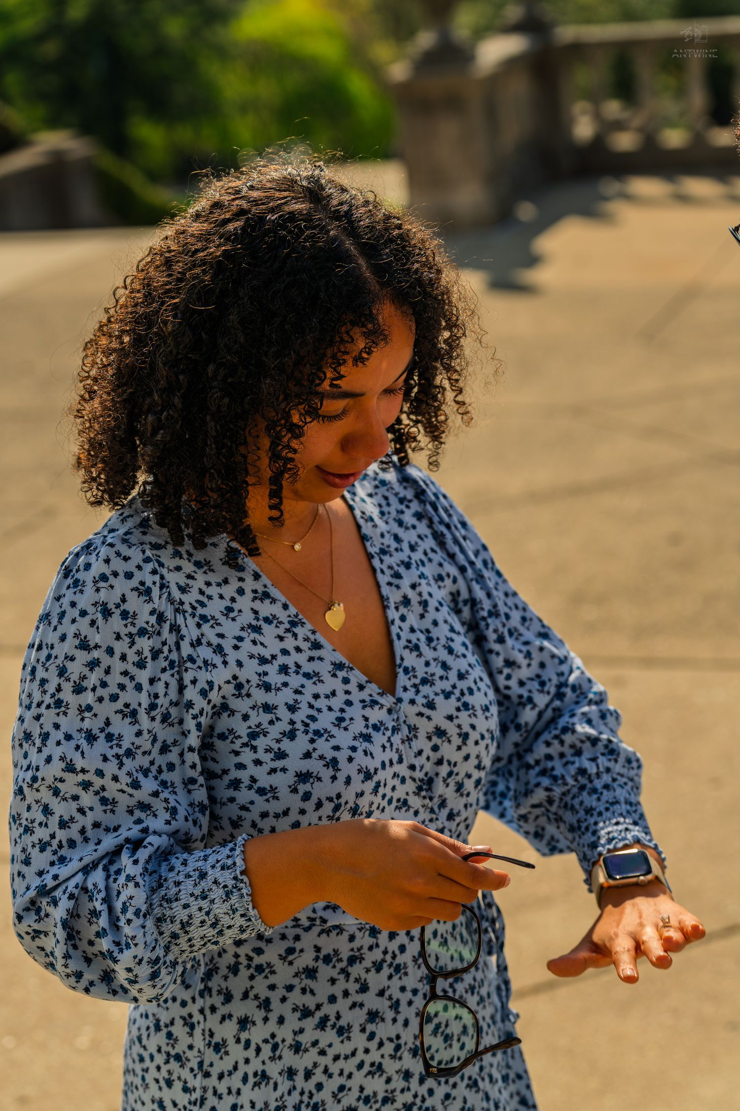 A curly-haired woman in a blue floral dress checks her phone while holding sunglasses outdoors in warm light.