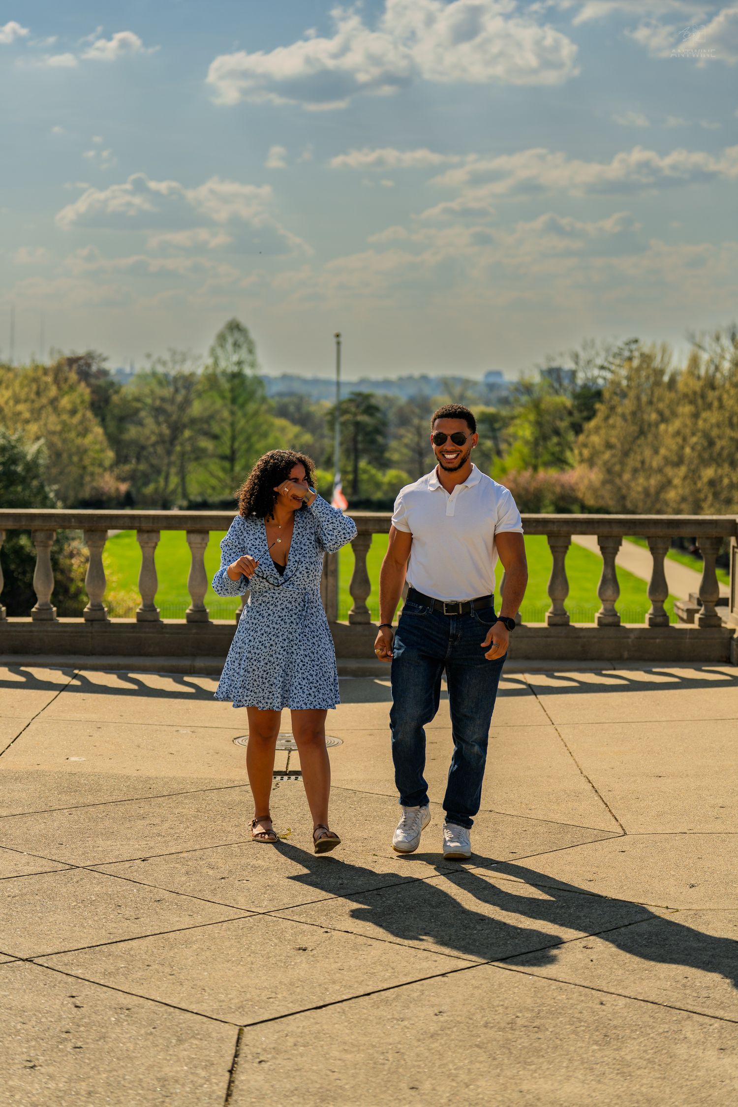 A stylish couple walks and laughs together on a sunlit stone terrace with park views behind.