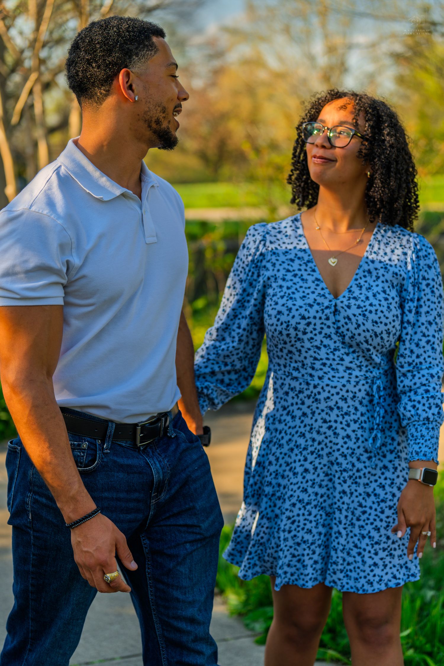 A couple gazes at each other lovingly while walking hand in hand along a sunlit park pathway.