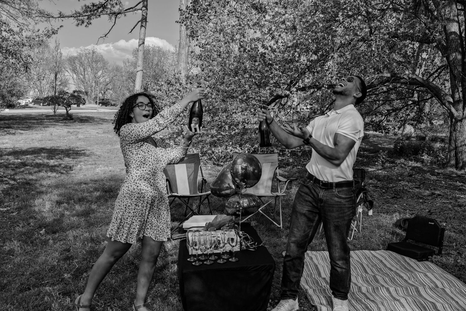 Black and white candid photo of a couple celebrating with champagne bottles at an outdoor park picnic.