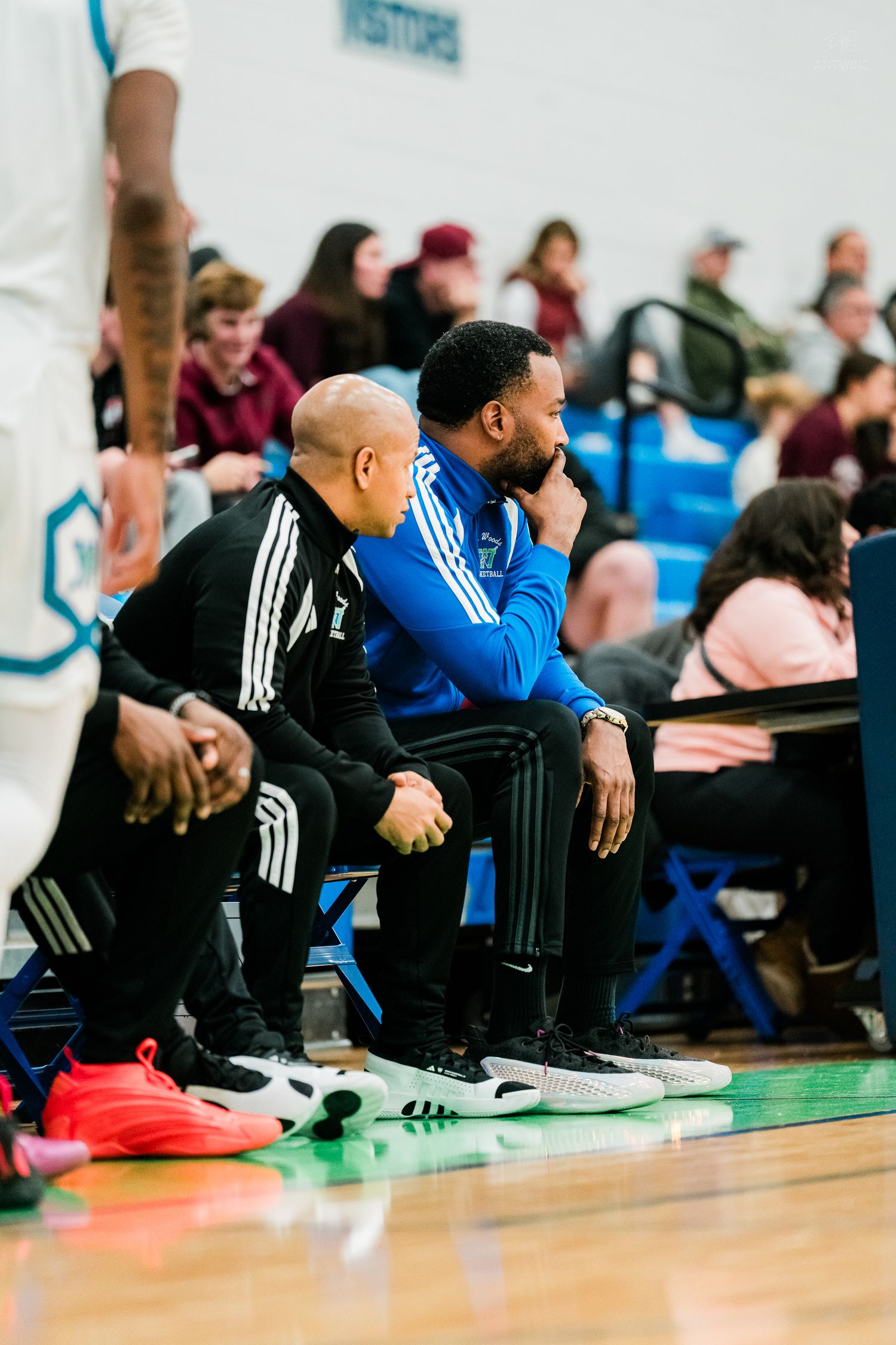 Two focused high school basketball coaches sit on the sideline bench watching gameplay in a packed gymnasium.