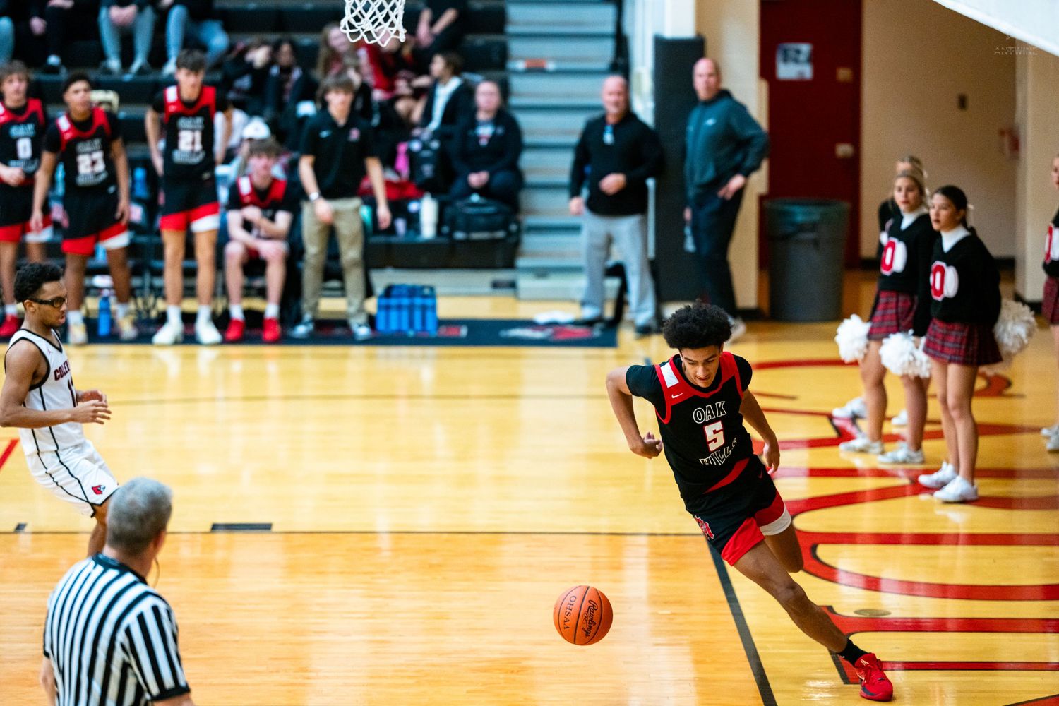 Player #5 in red uniform dribbles past referee during high school basketball game.