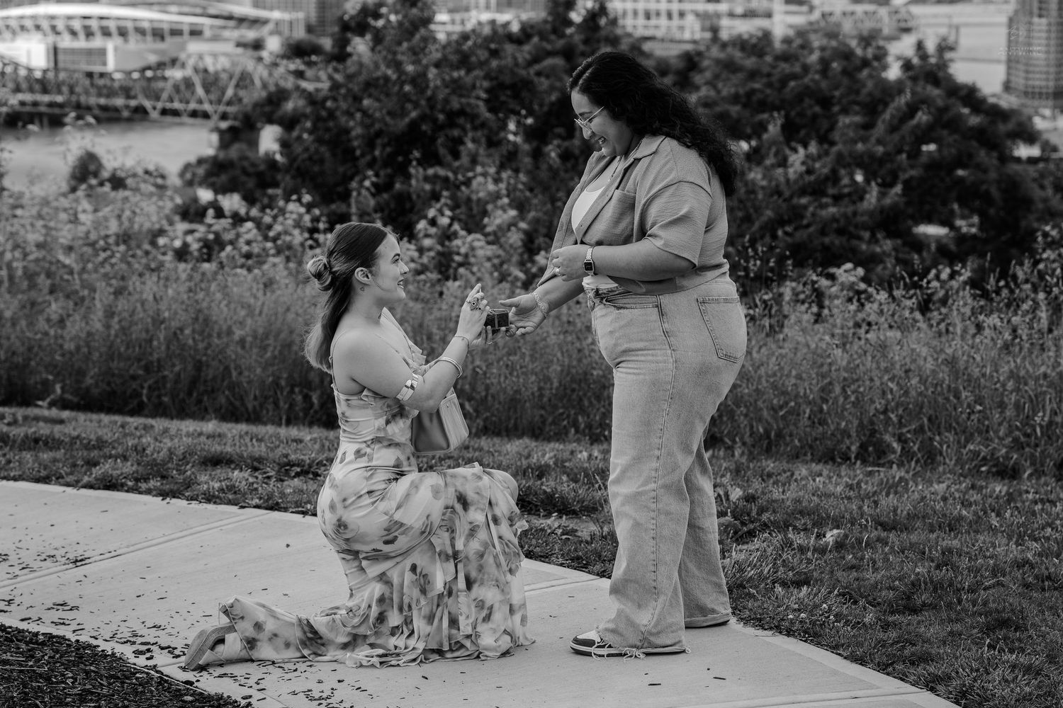 Black and white outdoor proposal photo of two women near urban greenery.