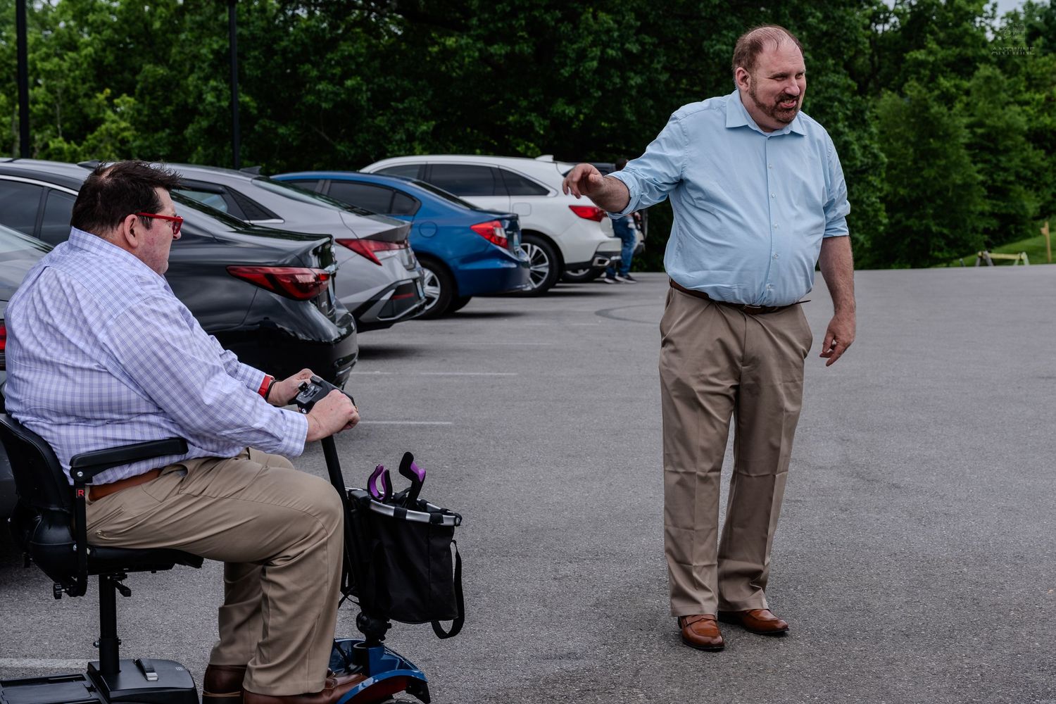 Two men in a parking lot, one on a mobility scooter as the other reaches out to greet him.