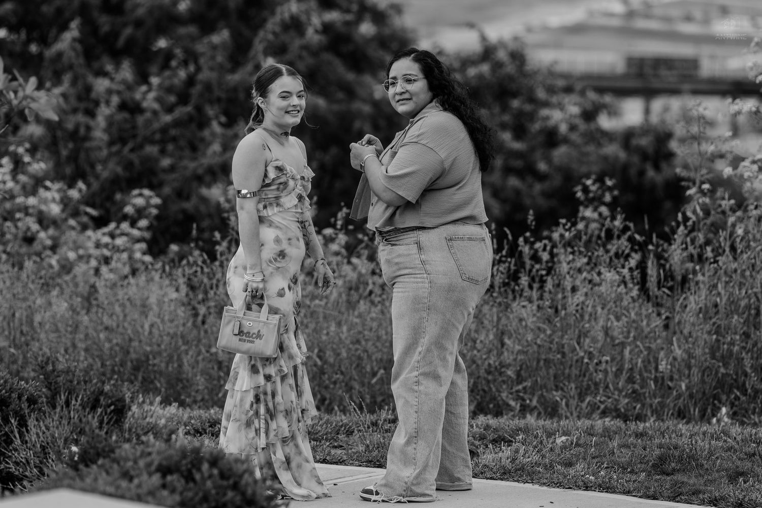 Two women pose together outdoors in a garden setting, one in a floral dress and one in casual attire.