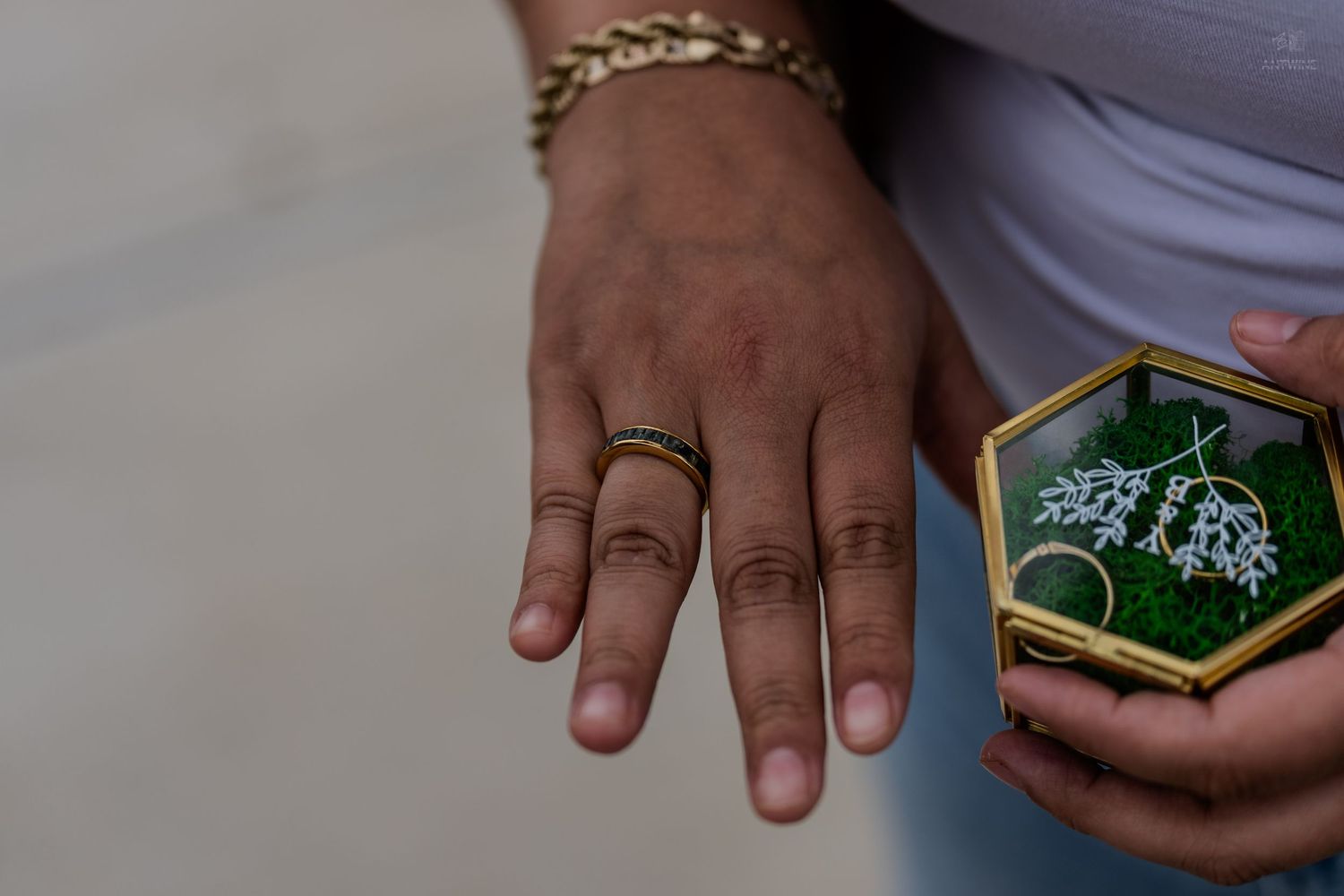 Hands displaying a gold wedding band beside a hexagonal glass ring box with green moss.