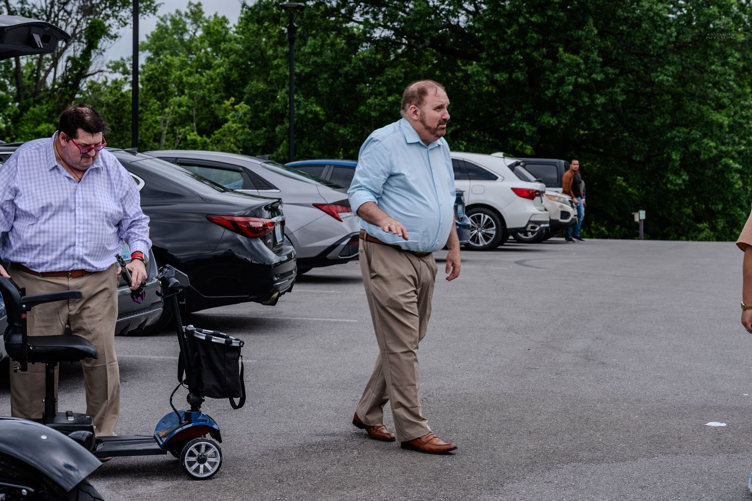Two men standing in a parking lot near a mobility scooter among parked cars.
